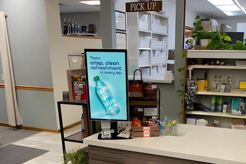 Countertop digital screen with a bottled-water ad at a pharmacy pickup counter