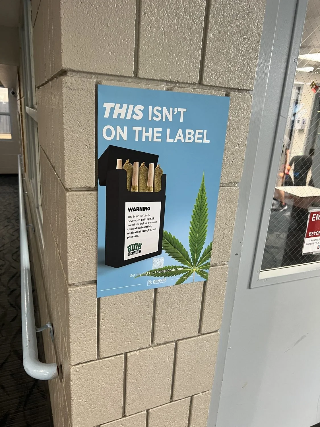 This Isn't On The Label cannabis-prevention poster on a cinder-block pillar in a school hallway, next to a door with a wired-glass window