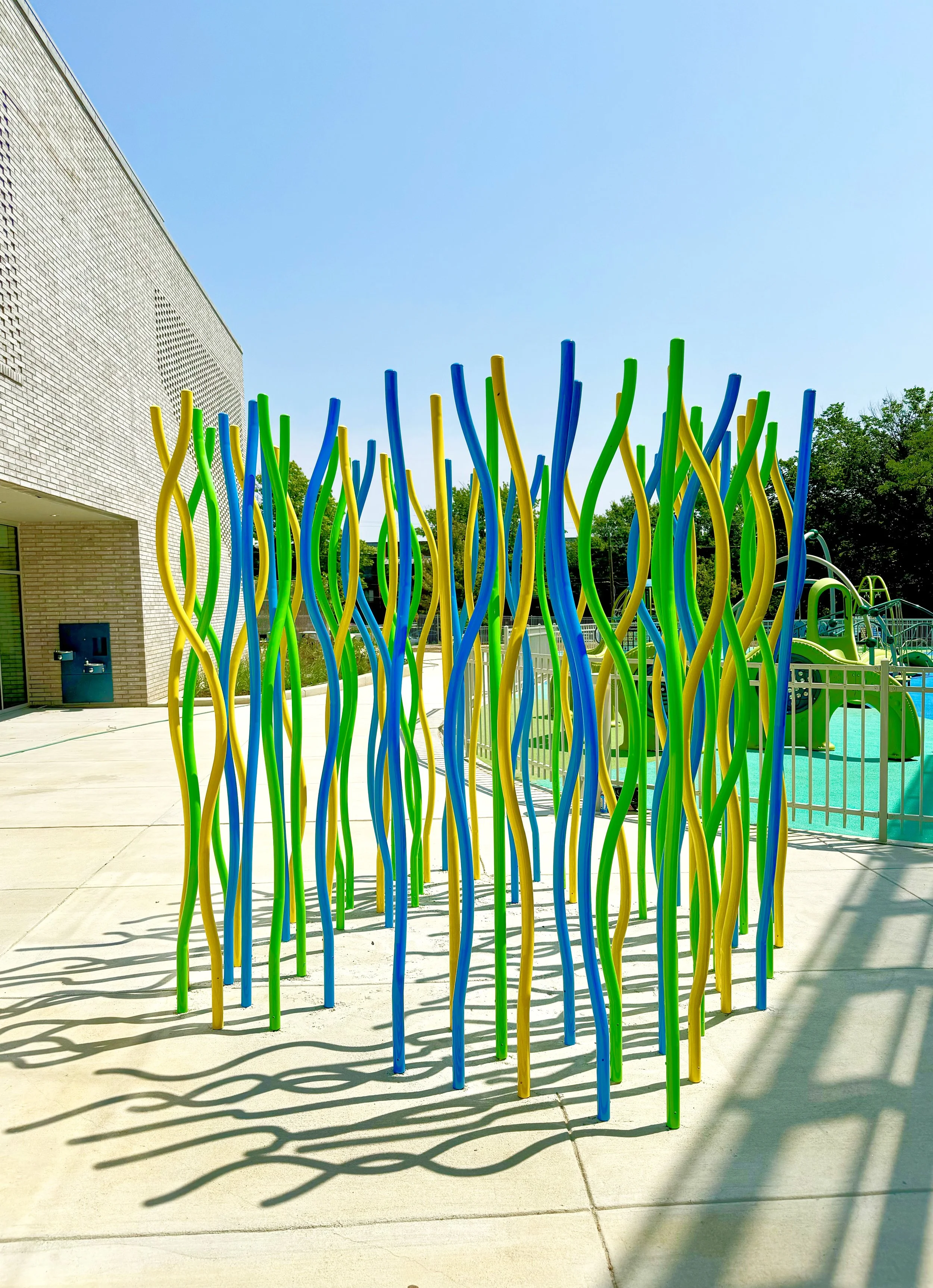 Colorful playground fence with green, yellow, and blue wavy metal poles casting shadows on the concrete ground outdoors, with a blue sky and trees in the background.