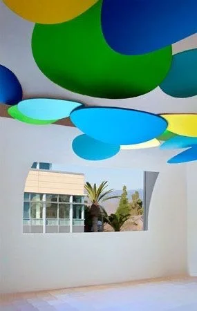 Colorful, round ceiling lights in blue, green, yellow, and teal on a white ceiling with a view of a modern building and palm trees through a window.