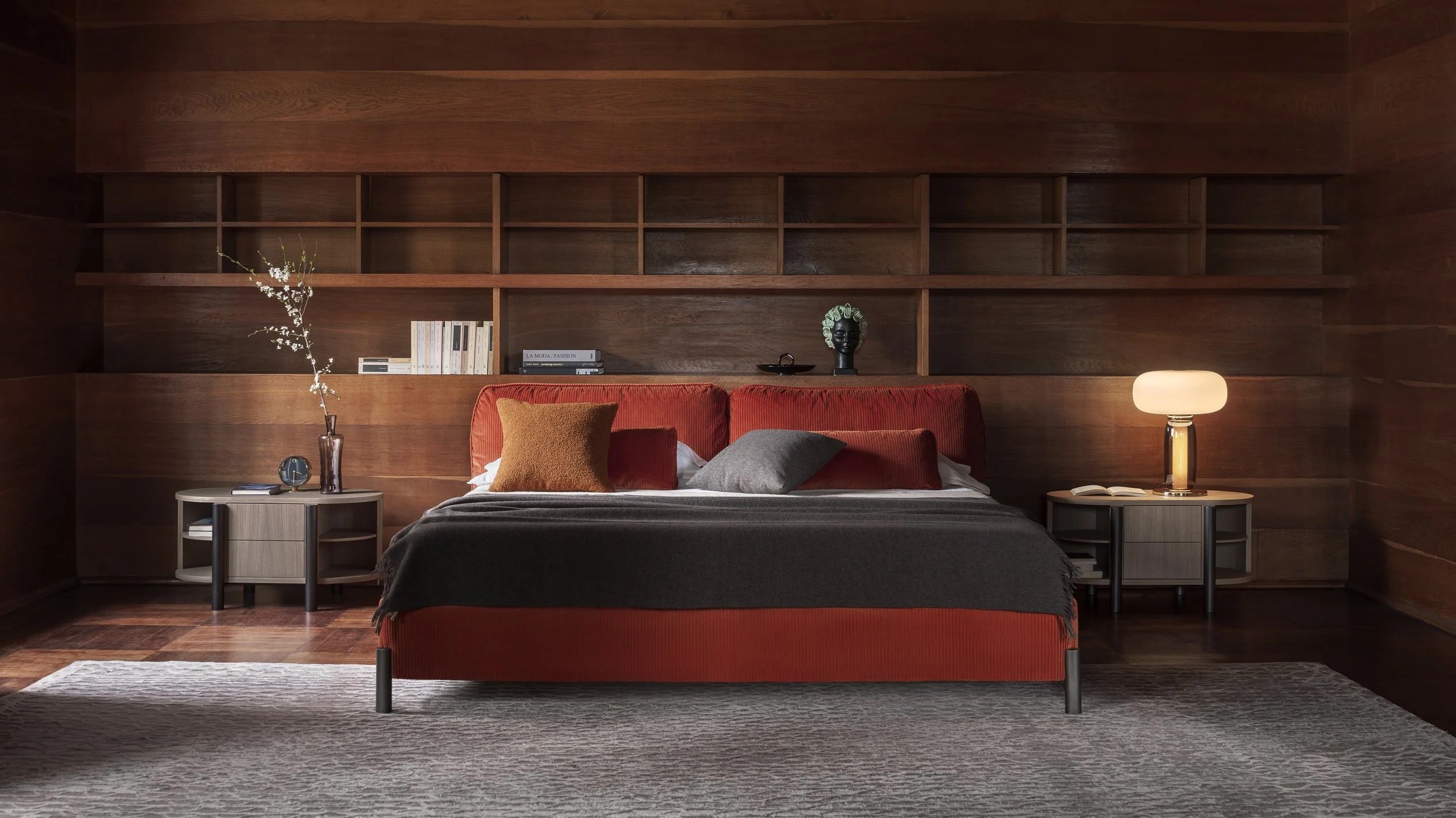 Bedroom with dark wood paneled walls, red upholstered bed with pillows, bedside tables with lamps, decorative items, and a rug.