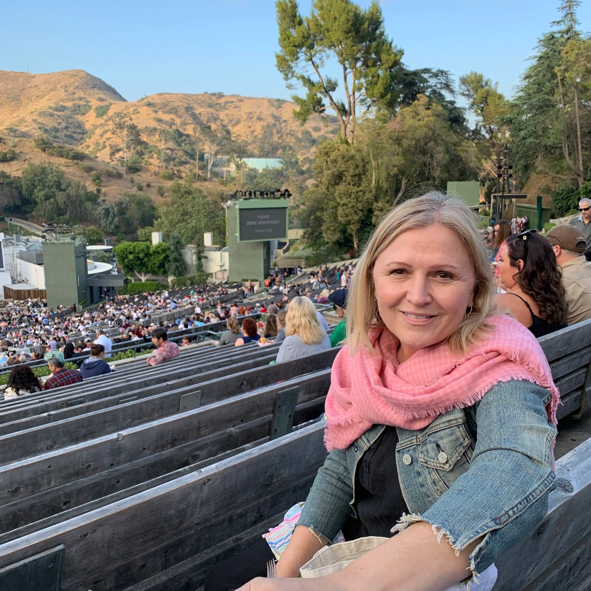 Barbara with blonde hair and a pink scarf over a denim jacket at an outdoor venue