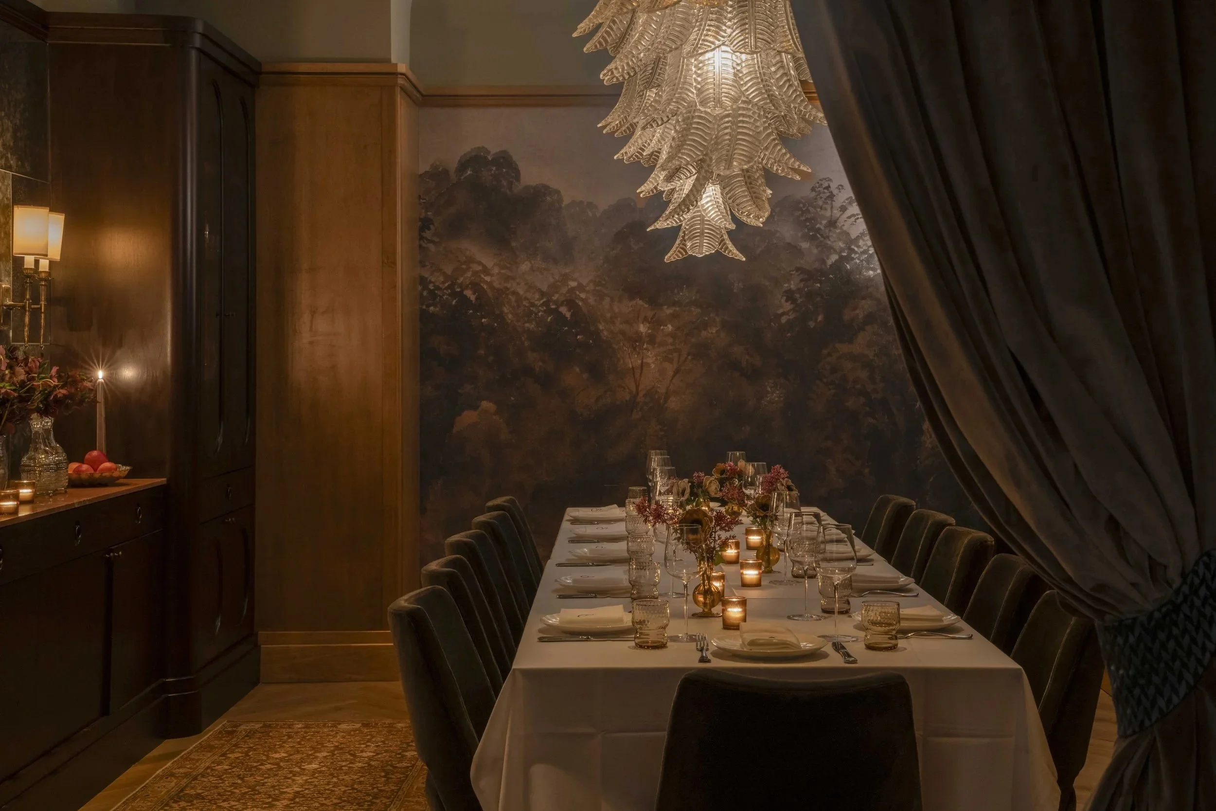 Elegant dining room with a long table set for a formal dinner, surrounded by dark velvet chairs, decorated with candles and floral centerpieces, in front of a large mural of a forest scene, with a luminous leaf-shaped chandelier hanging above, partially concealed by dark curtains.