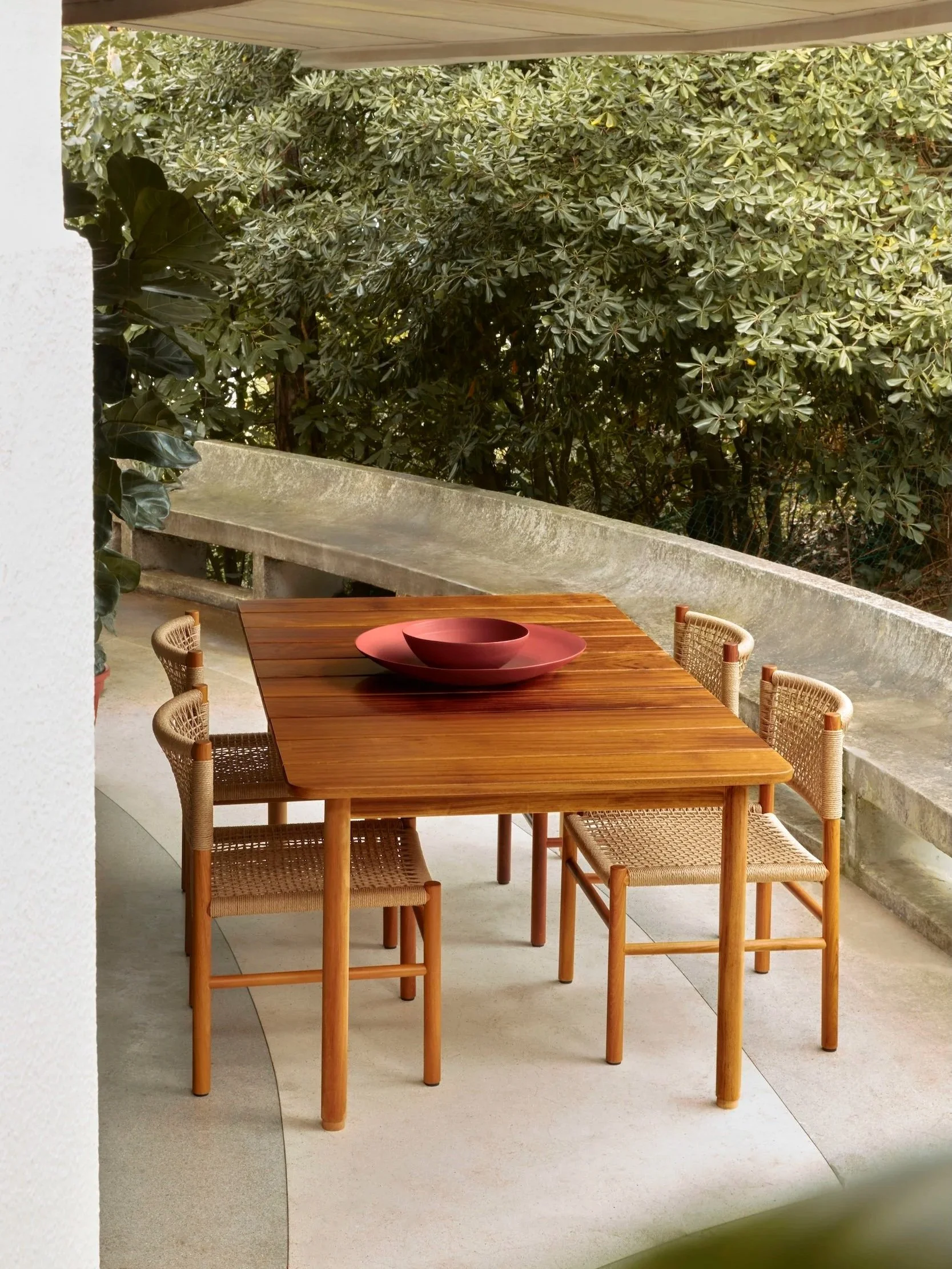 A wooden outdoor dining table with four wicker chairs and a pair of nested red dishes on top, set on a patio surrounded by greenery.