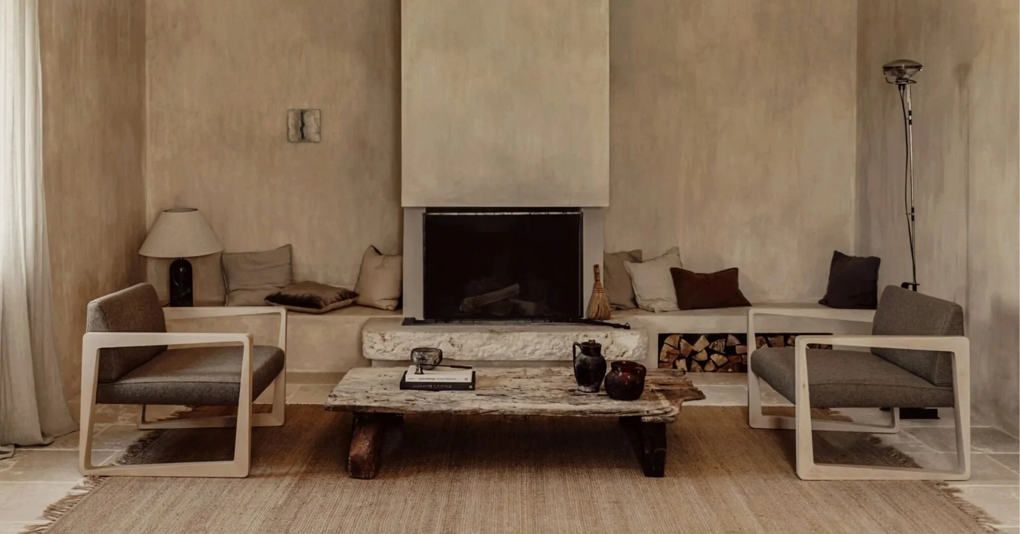 Living room with a rustic wooden coffee table, beige walls, a fireplace, gray and beige lounge chairs, and decorative cushions, with a floor lamp on the right side.