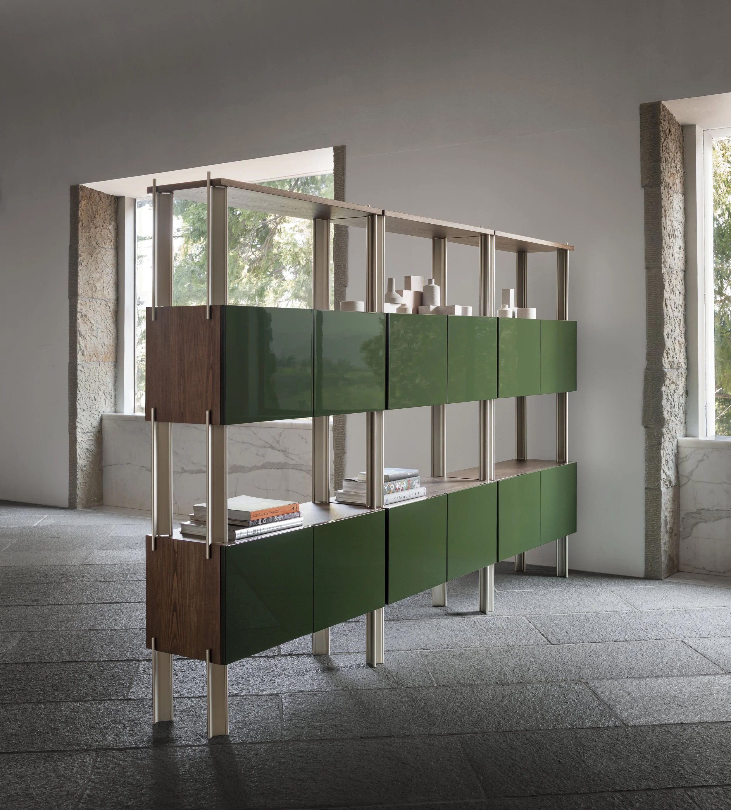 A modern bookshelf with green and wood panels in a room with stone window frames and large windows letting in natural light.