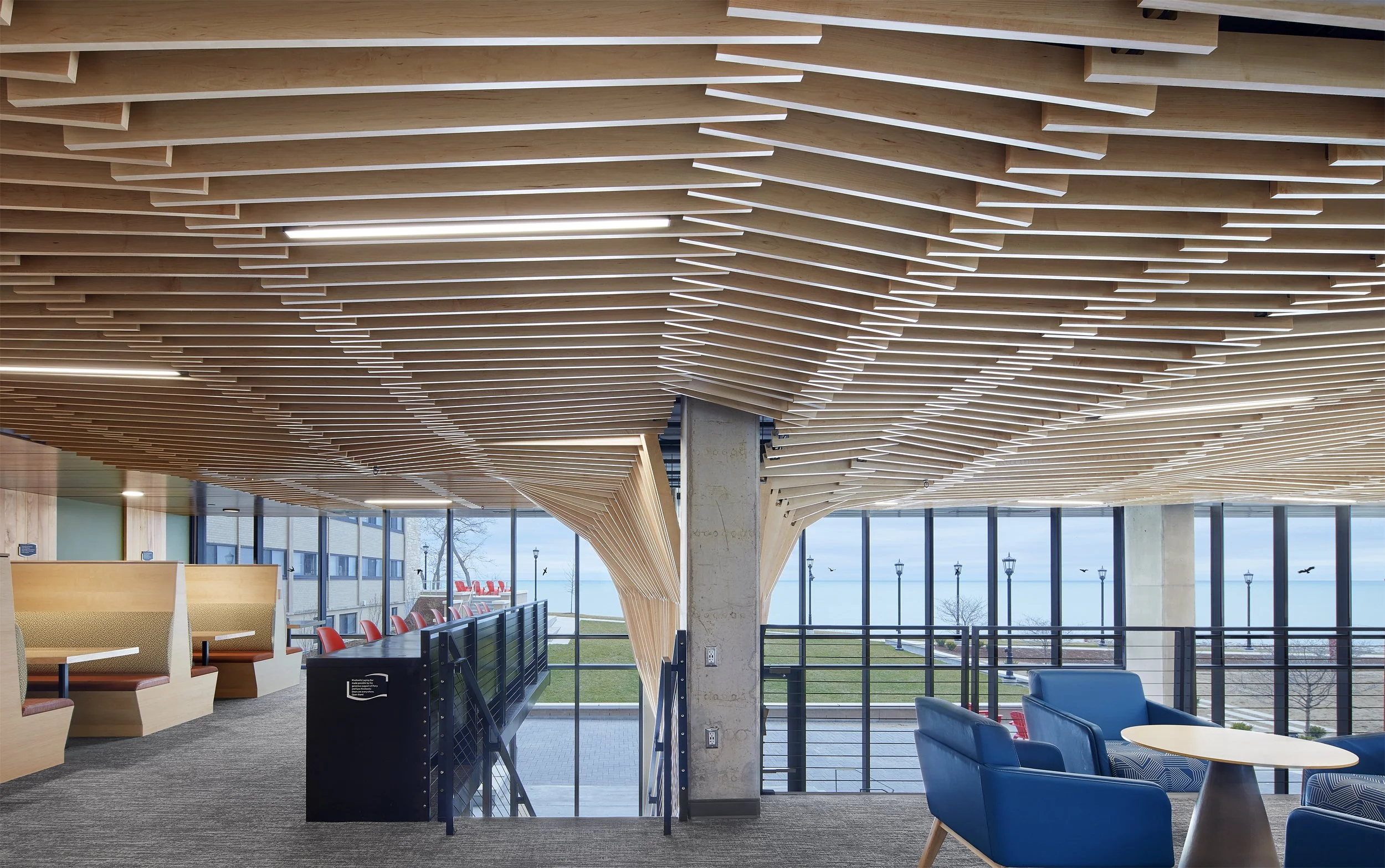 Interior of a modern building with a ceiling made of layered wooden slats, large windows showing an outdoor view of a body of water and a grassy area, and seating areas with blue chairs and booth-style seats.