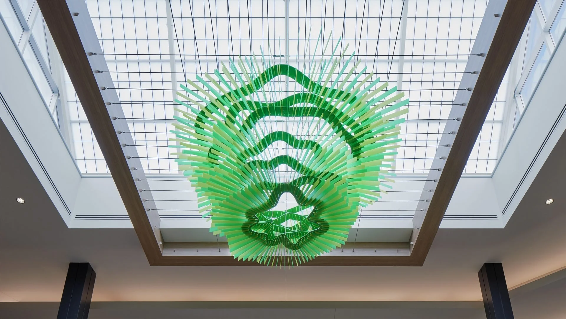 Green and white abstract statement chandelier hanging from a skylight ceiling in a modern building interior.