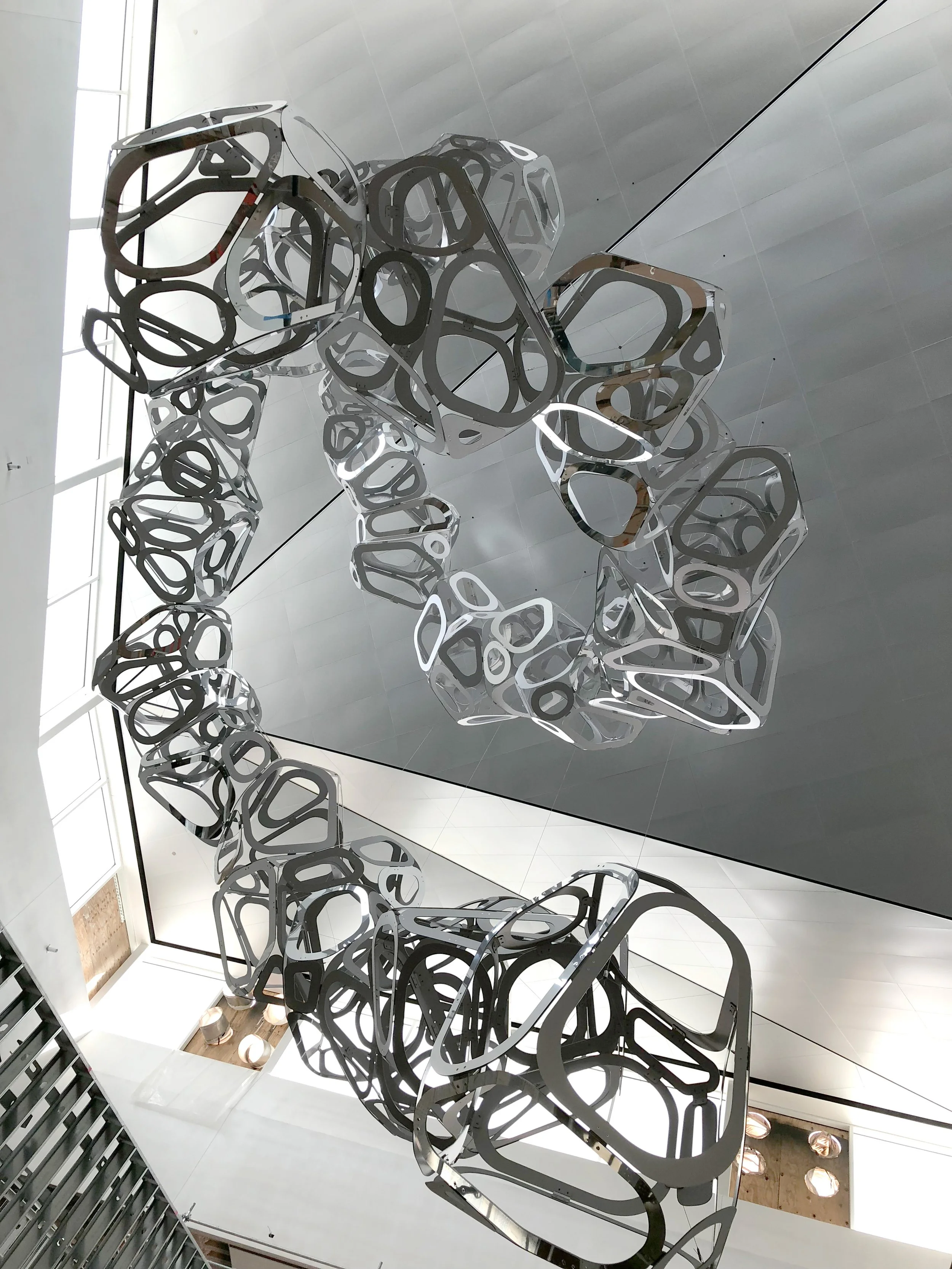 Modern abstract metal sculpture installation hanging from the ceiling in a bright, spacious interior.