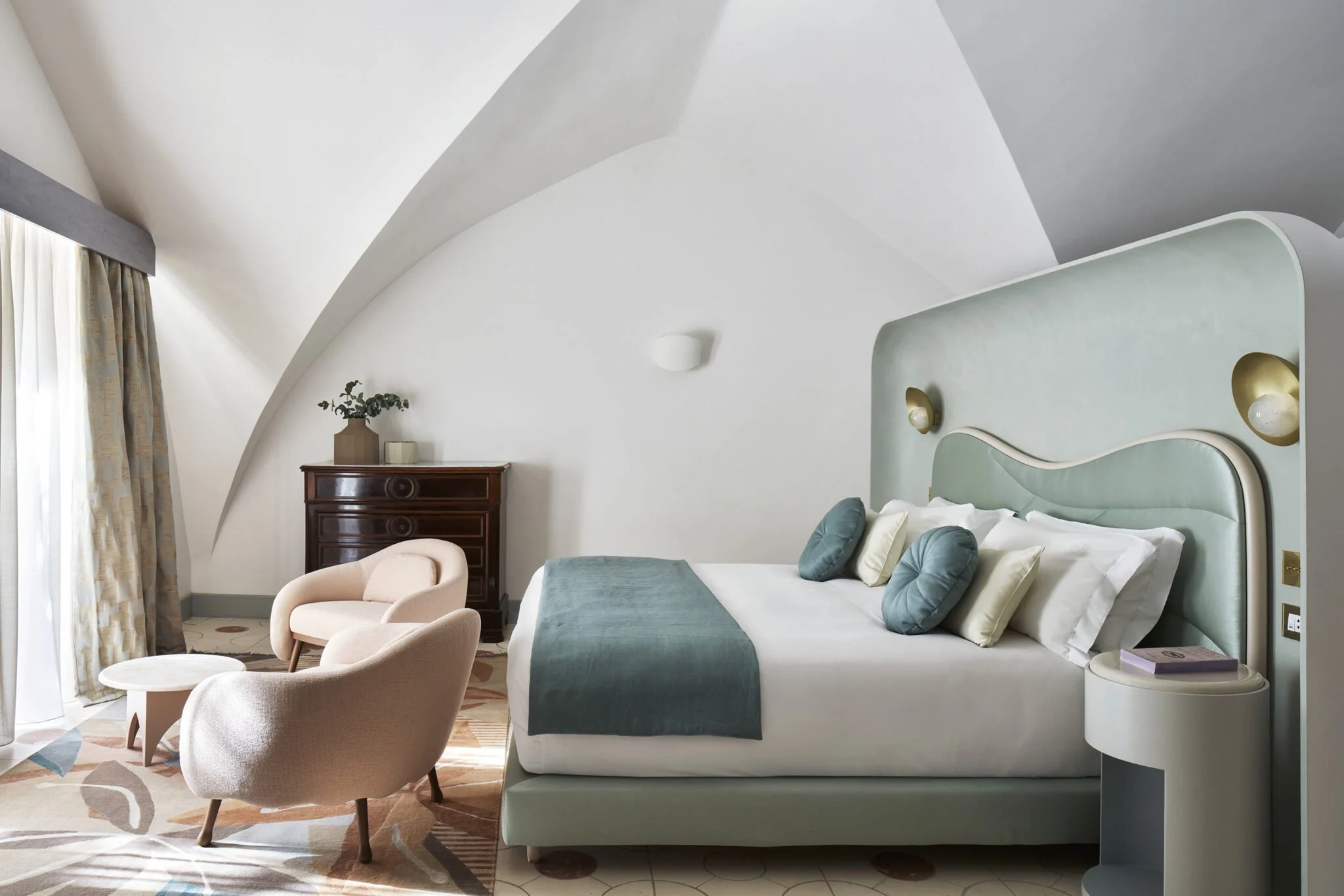 A modern  room with a large bed, a pastel-colored headboard, circular pillows, and a textured blanket. Beside the bed is a curved white nightstand with a book and a light. In the background, there are two beige armchairs and a small table, a wooden dresser with plants and decorations, and curtains covering a window or balcony door.