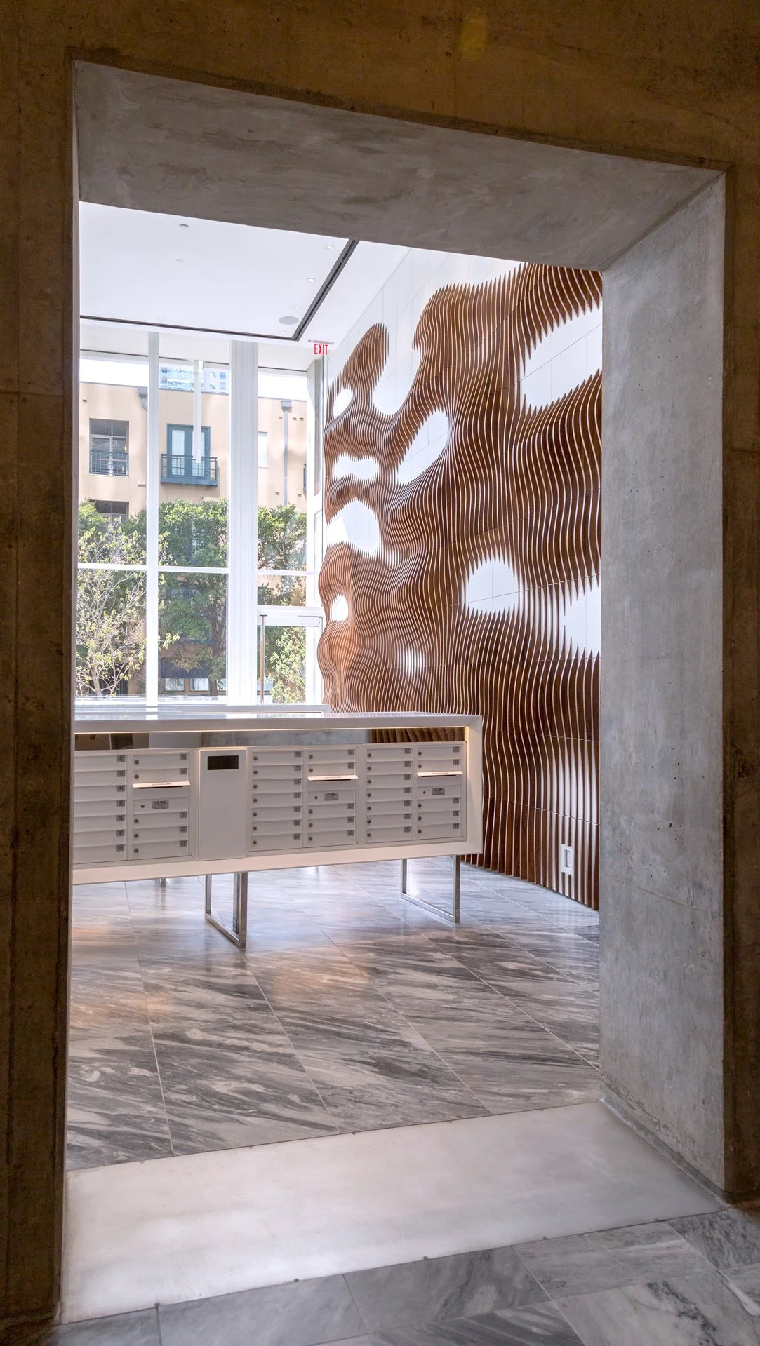 Interior view of an apartment building lobby with a wall made of wooden slats with circular cutouts, a row of white mailboxes, marble flooring, and large glass windows showing trees and neighboring buildings outside.