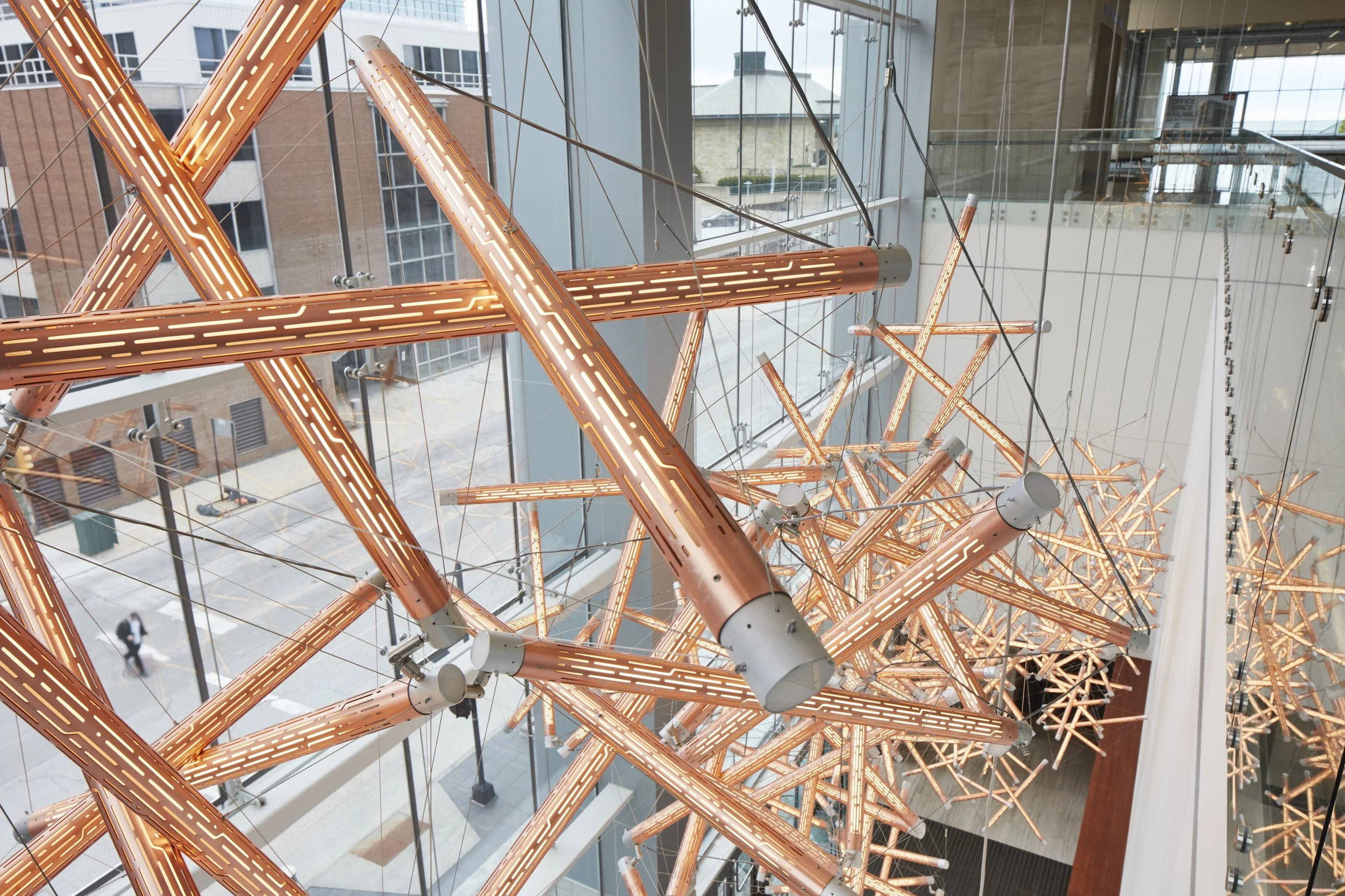 Interior view of a modern building with large glass windows and a striking artistic light fixture made of numerous interconnected copper pipes with integrated lighting, hanging from the ceiling.