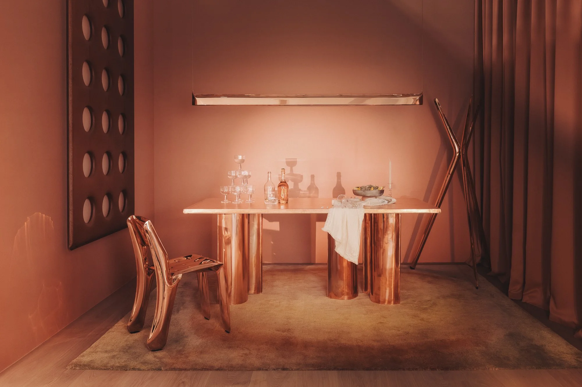 A room with a pink wall and copper-colored furniture. There is a table with glassware and bottles, a chair, and a curtain on the right side.