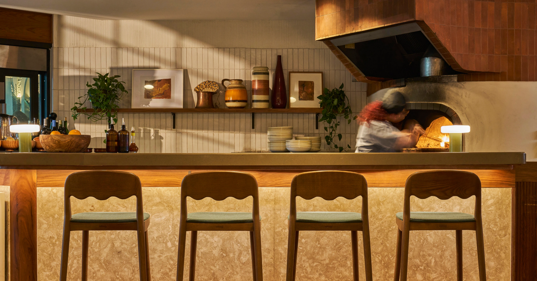 Interior of a restaurant or cafe with a wooden bar counter and three wooden chairs in the foreground. A chef is working in a wood-fired oven in the background. Decor includes potted plants, framed artwork, and decorative vases on a shelf.