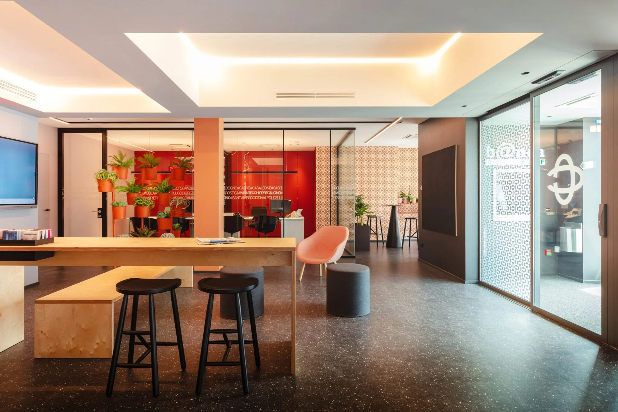 Modern office reception area with a wooden desk, pink armchair, black and gray stools, potted plants, and glass-walled meeting room with red decor.