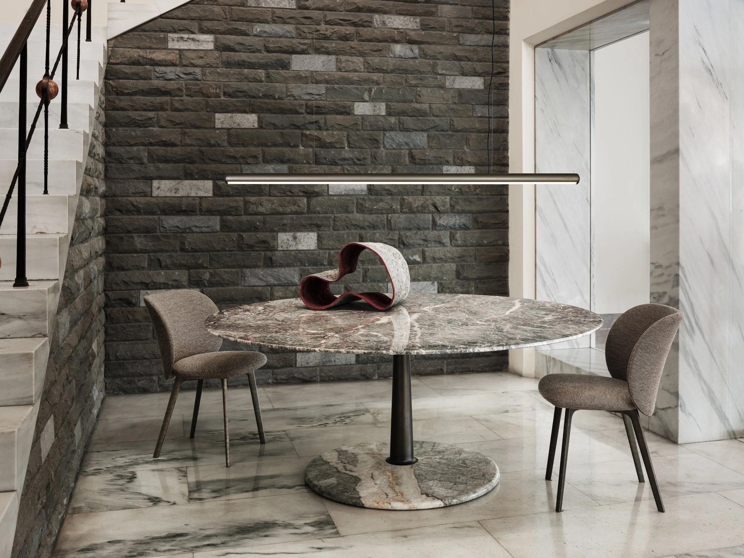 A round marble table with a decorative sculpture on top, two upholstered chairs, a brick wall, and marble flooring in an interior space.