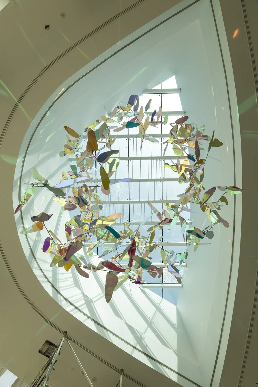 Colorful abstract sculpture hanging from a ceiling inside a modern building with large windows and a skylight.