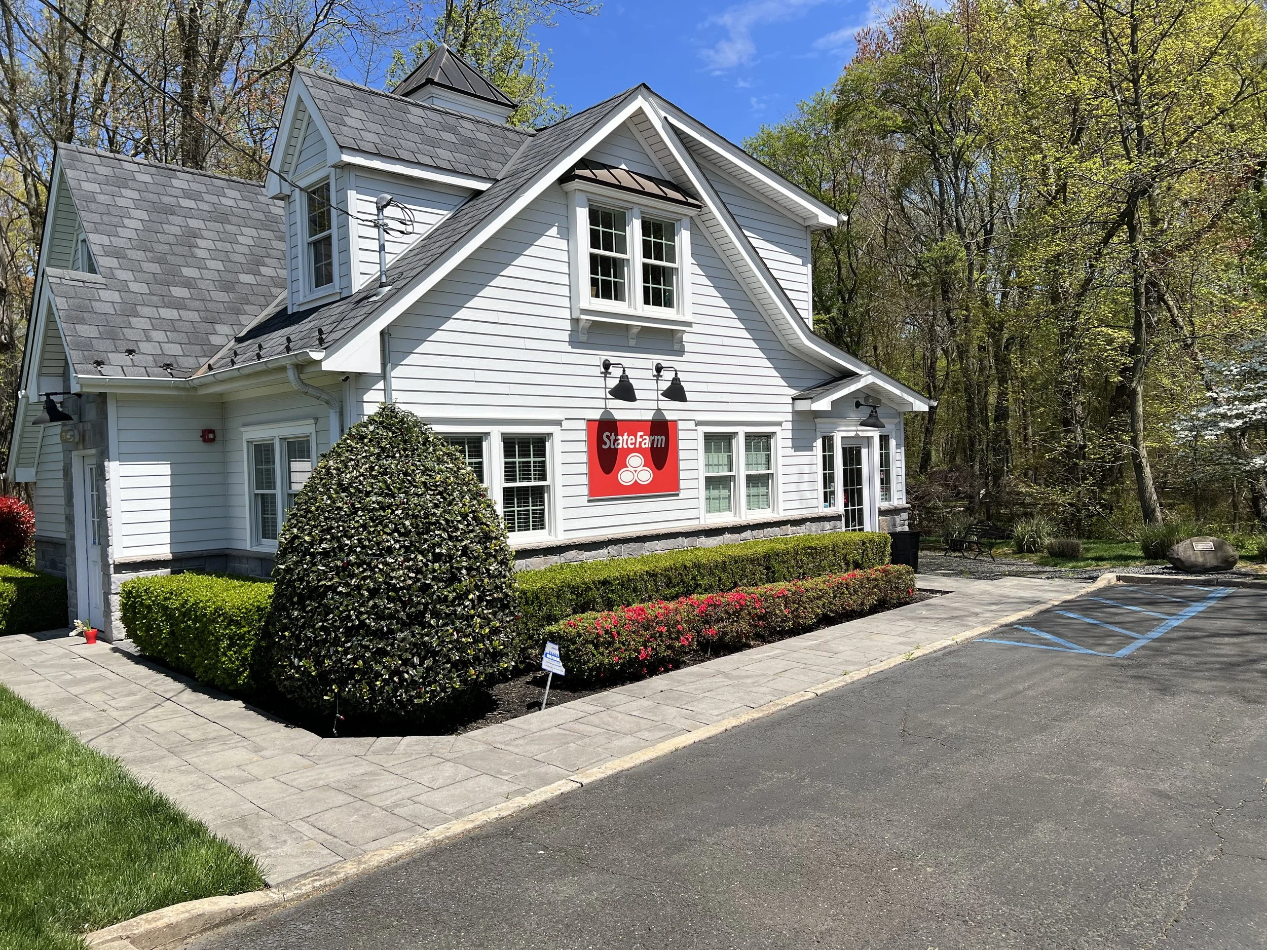 A white two-story house with a small tower, surrounded by trees, displaying a State Farm insurance sign on its front. The house has a well-maintained garden with hedges and flowers, and there is a parking lot with spaces marked for disabled parking in front.