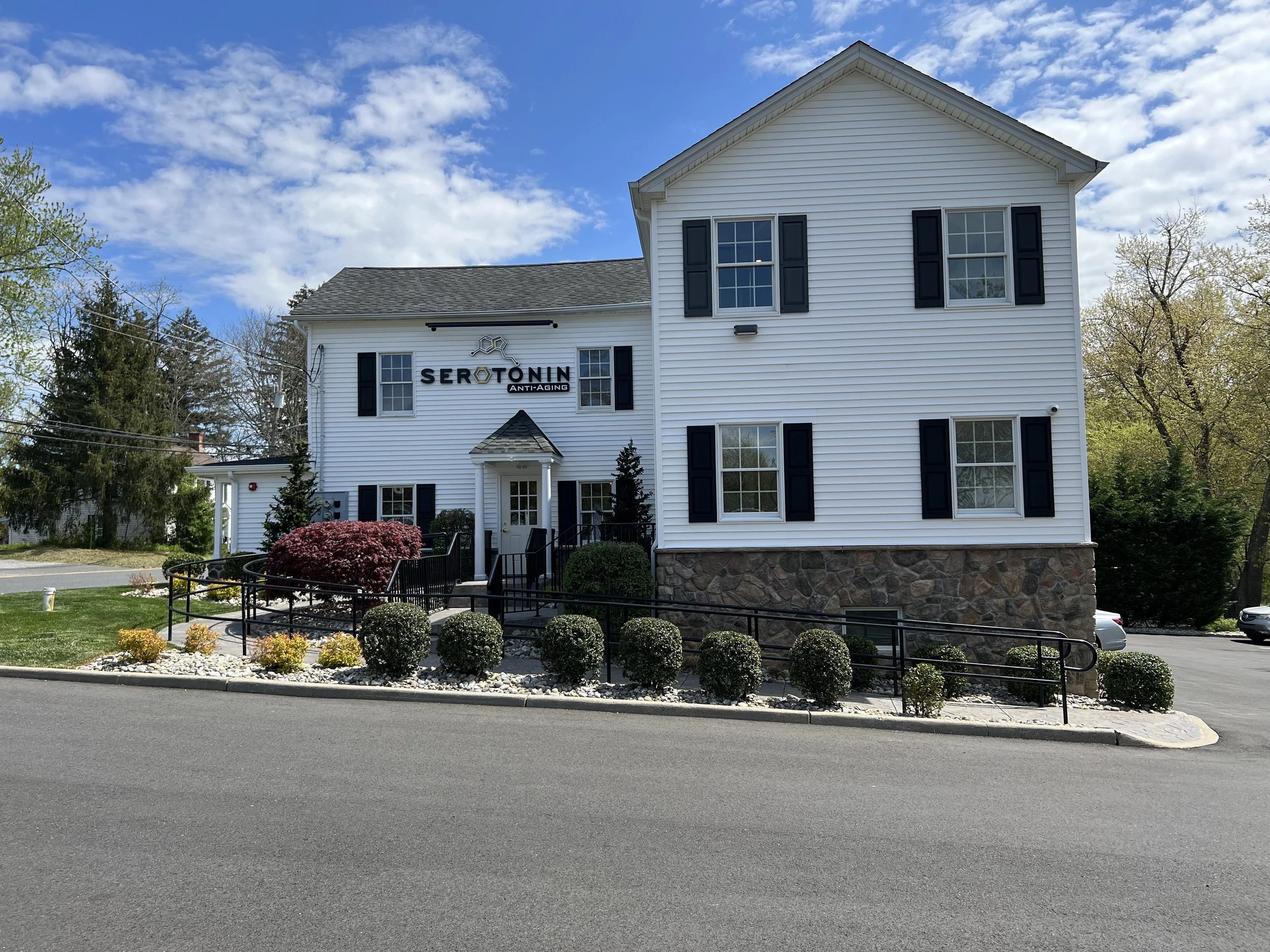 A white multi-story building with black shutters and a sign that reads 'Serotonin Anti-Aging'. The building has landscaping with bushes, small trees, and decorative rocks, and a parking lot is visible. Blue sky with clouds is overhead.