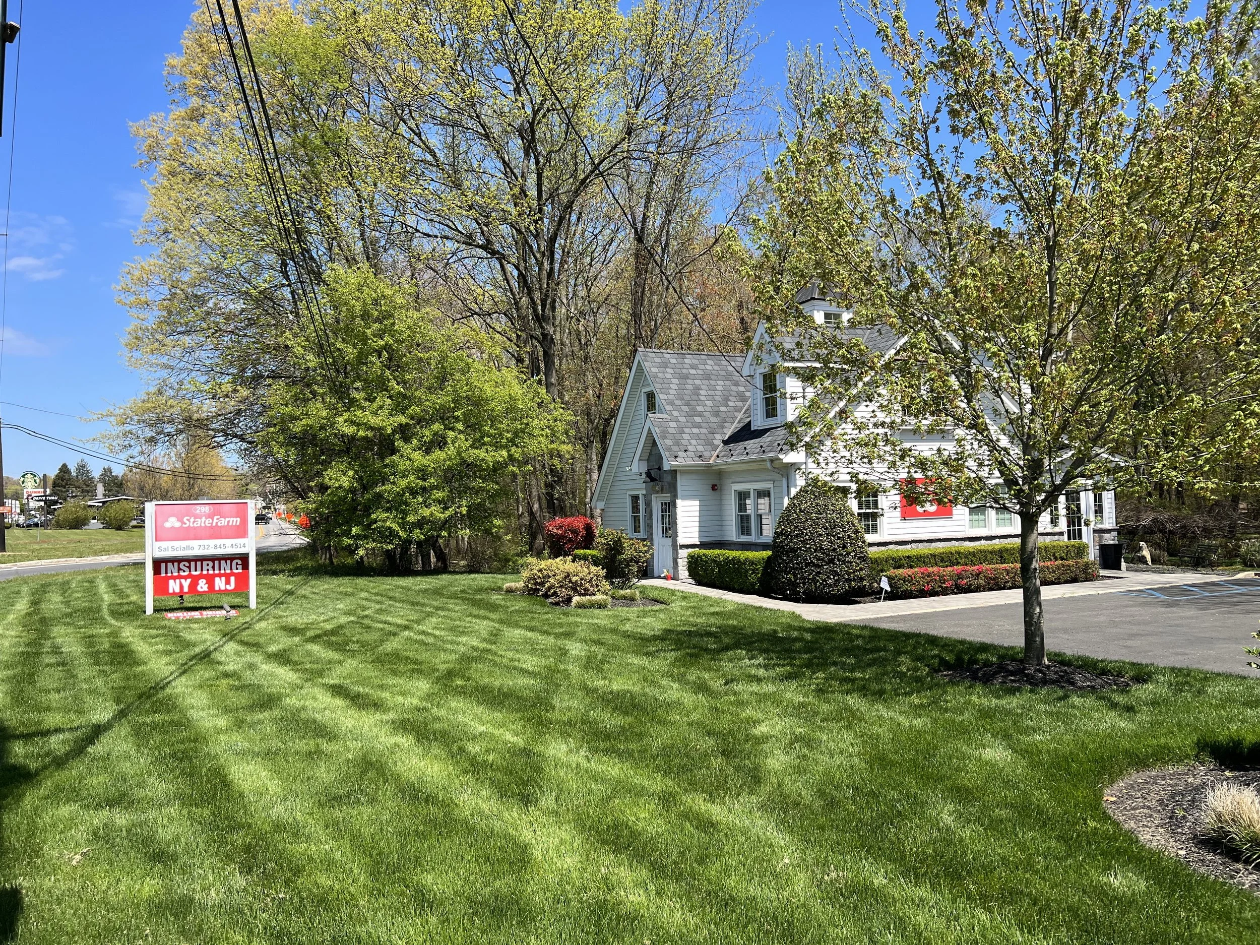A well-maintained lawn with a small tree, bushes, and a white house with gray roof shingles, surrounded by flowering plants and trees, under a bright blue sky.