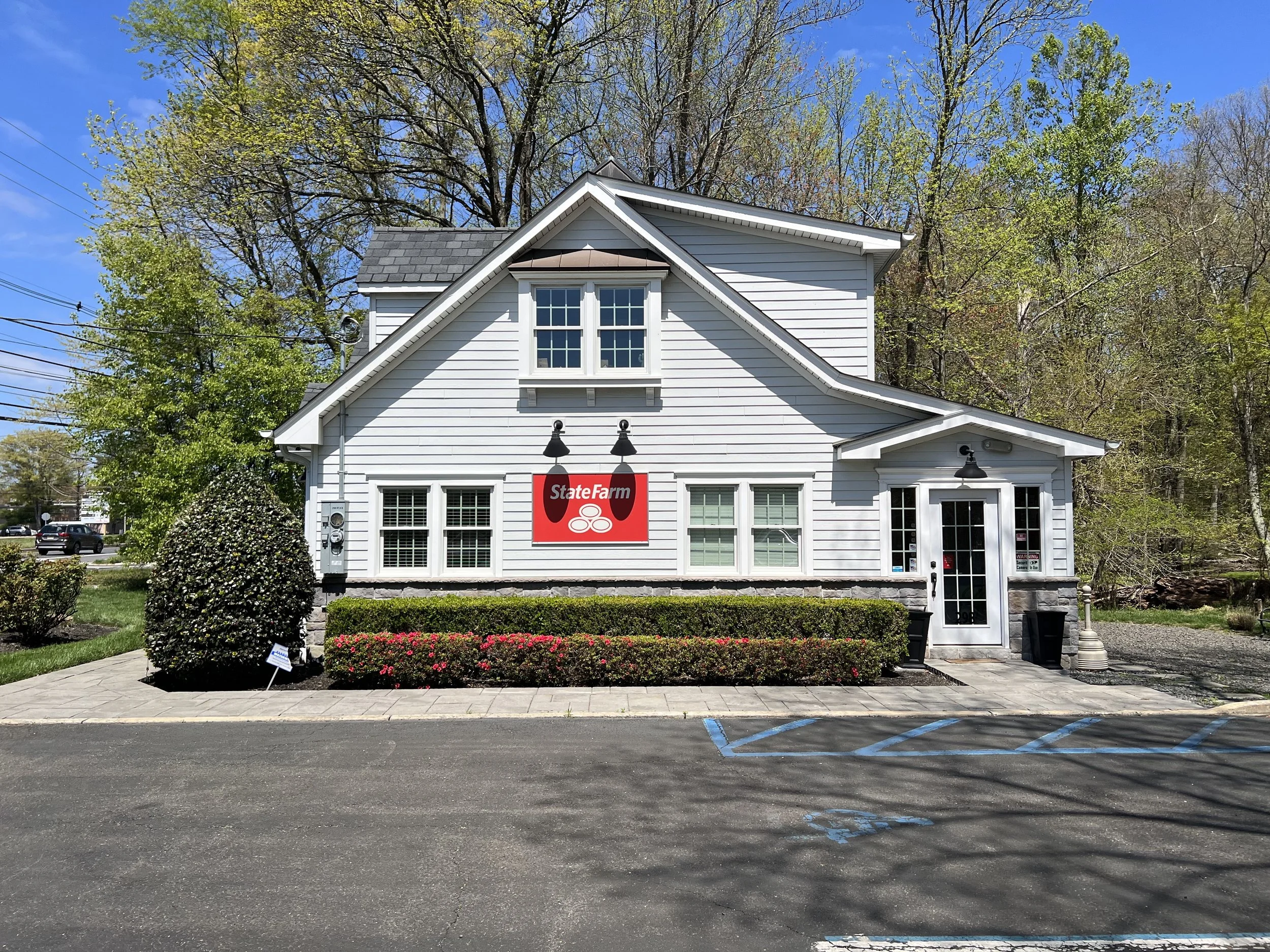 A white building with a sloped roof and a  sign for State Farm insurance on its front. The building has green bushes and pink flowers in front, with parking spaces and a parking lot in the foreground, and trees in the background.