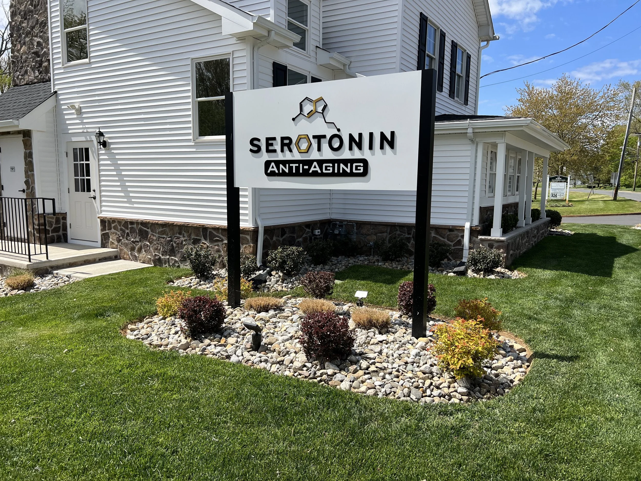 Sign for Serotonin Anti-Aging clinic in front of a white house with a stone foundation, surrounded by a green lawn and landscaped with rocks and small bushes.