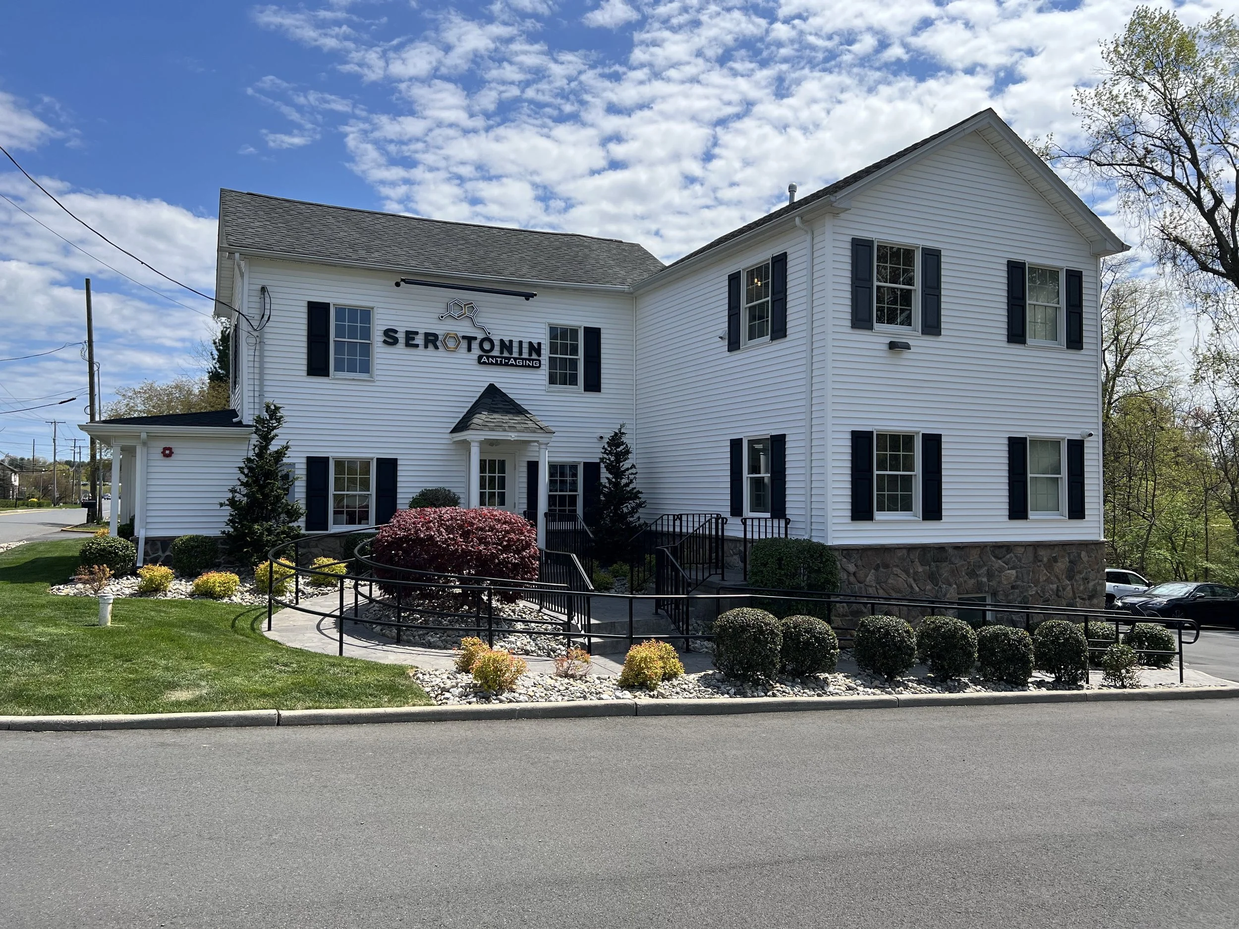 White two-story building with black shutters and a sign that reads 'Serotonin Anti-Aging' on the front, surrounded by neatly landscaped bushes and flowers, under a partly cloudy sky.