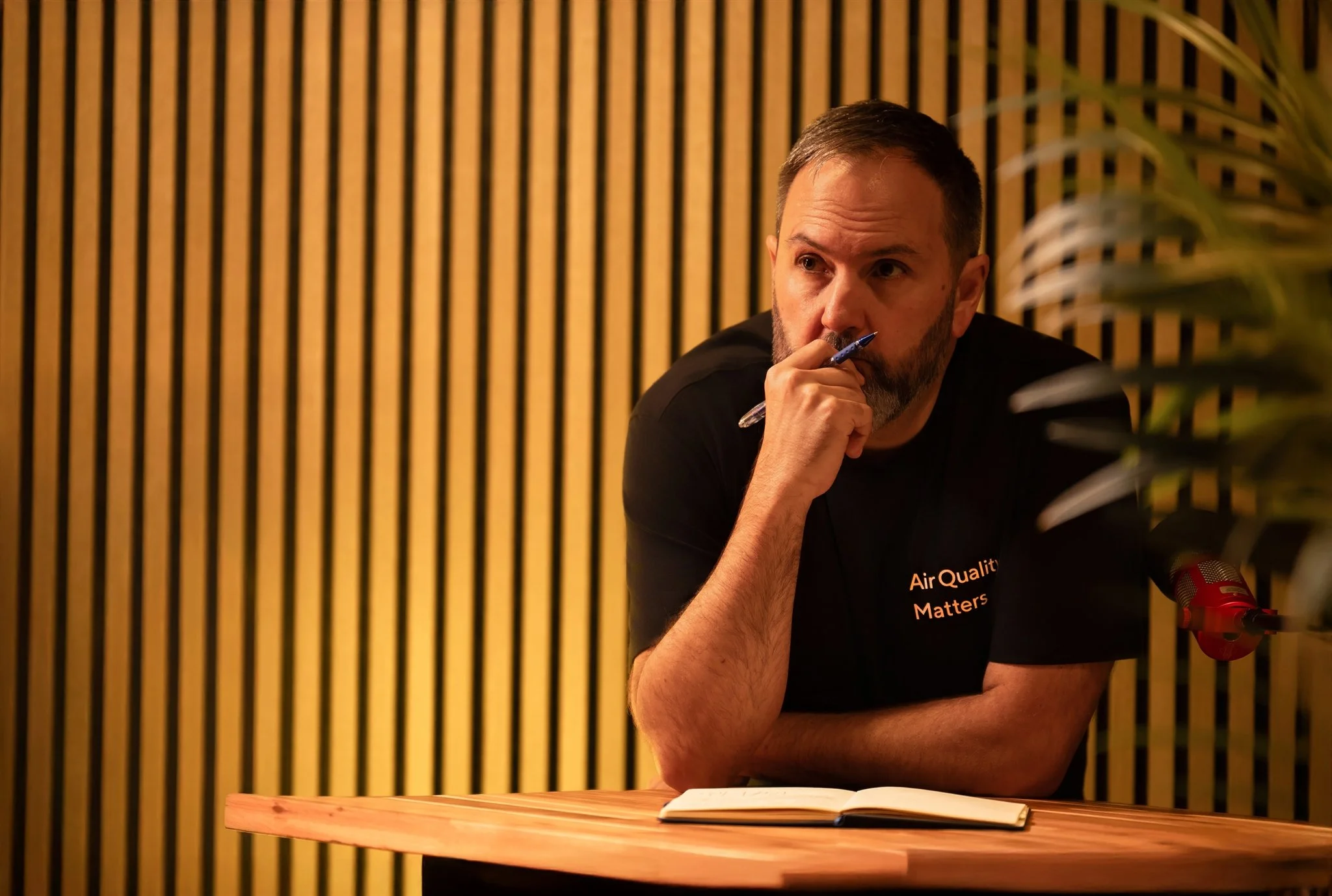 Cover image for a podcast celebrating one year of episodes, featuring a man with a beard in a black t-shirt crossing his arms, dark background with a crowd and a bright light source, and text about podcasts, air quality, and more.