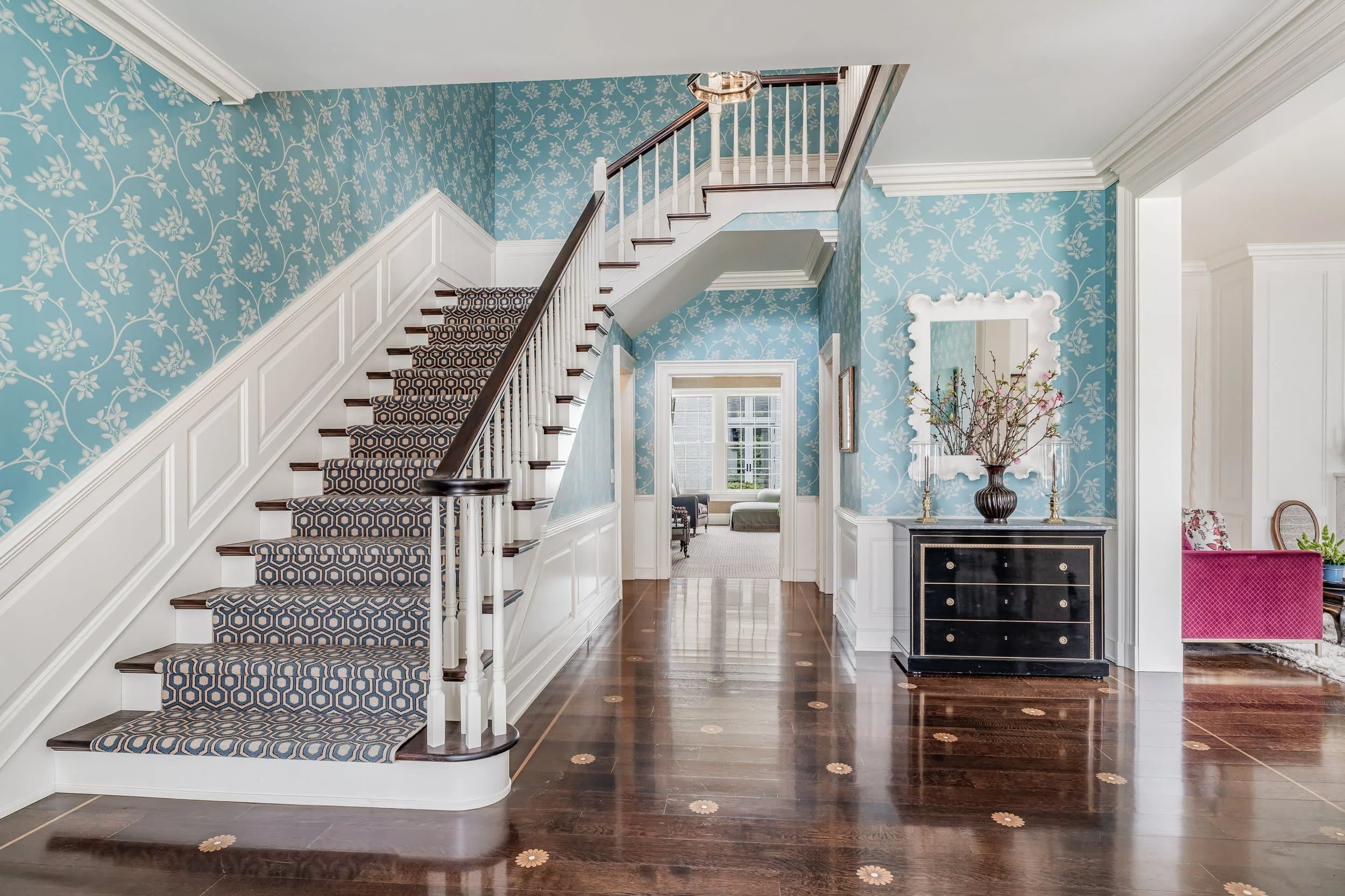 Bright entryway perfect for real estate, featuring patterned blue wallpaper, wooden stairs with runner, and a black console table with mirror.