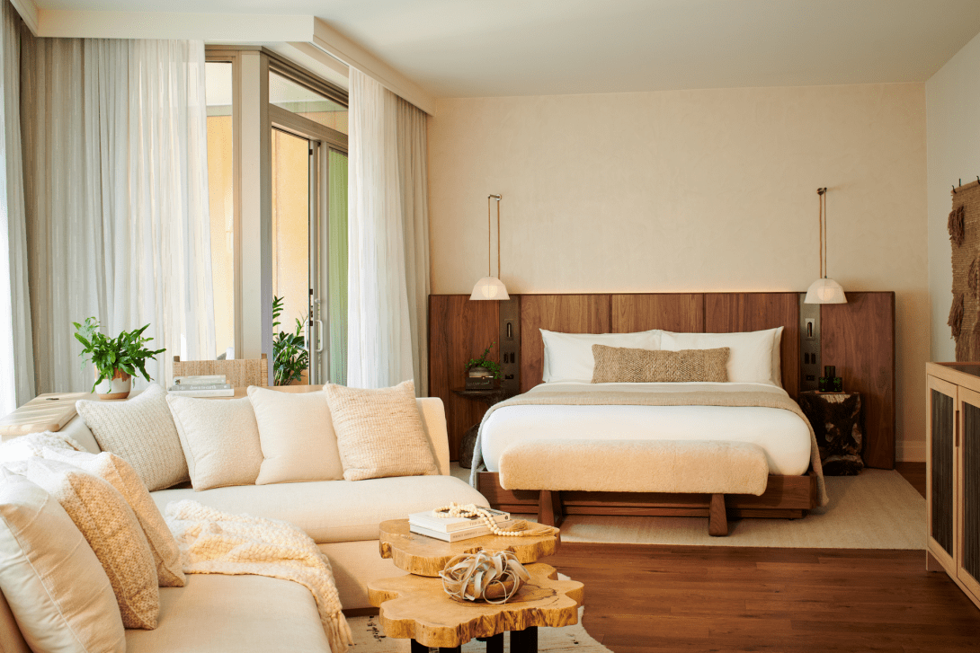 Modern bedroom with a large bed, white bedding, wooden headboard, beige pillows, and hanging white pendant lamps. There is a cream sofa with matching pillows and a rustic wooden coffee table in front. Large glass doors with sheer curtains lead outside to a patio with potted plants.