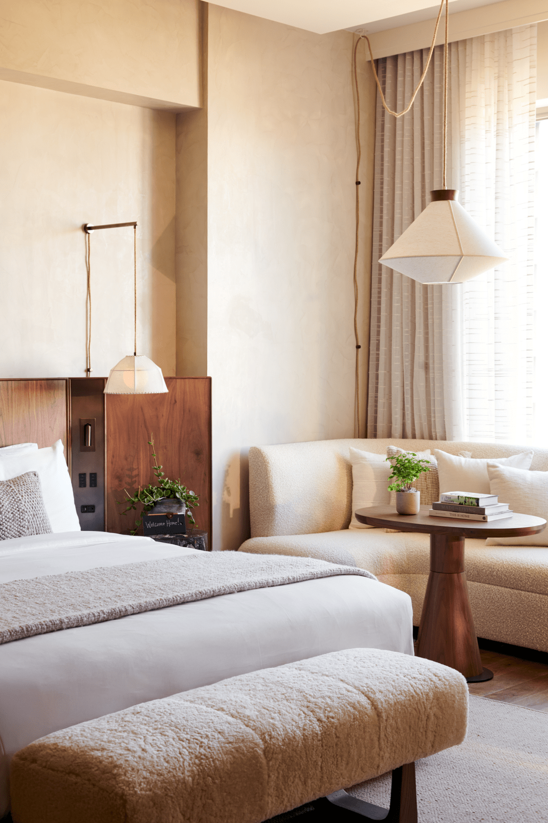 A hotel room with a bed, a beige couch, a round wooden table with a green potted plant and books, and a window with curtains. There are two hanging pendant lights and a small potted plant on a nightstand.