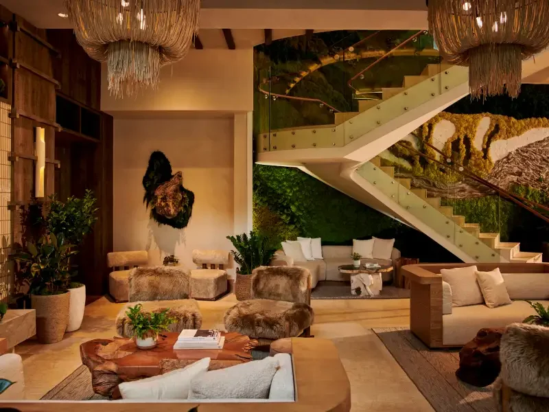 Luxurious living room with fur-covered chairs, a beige sofa, potted plants, and wall art; staircase with green moss wall backdrop and decorative chandeliers.