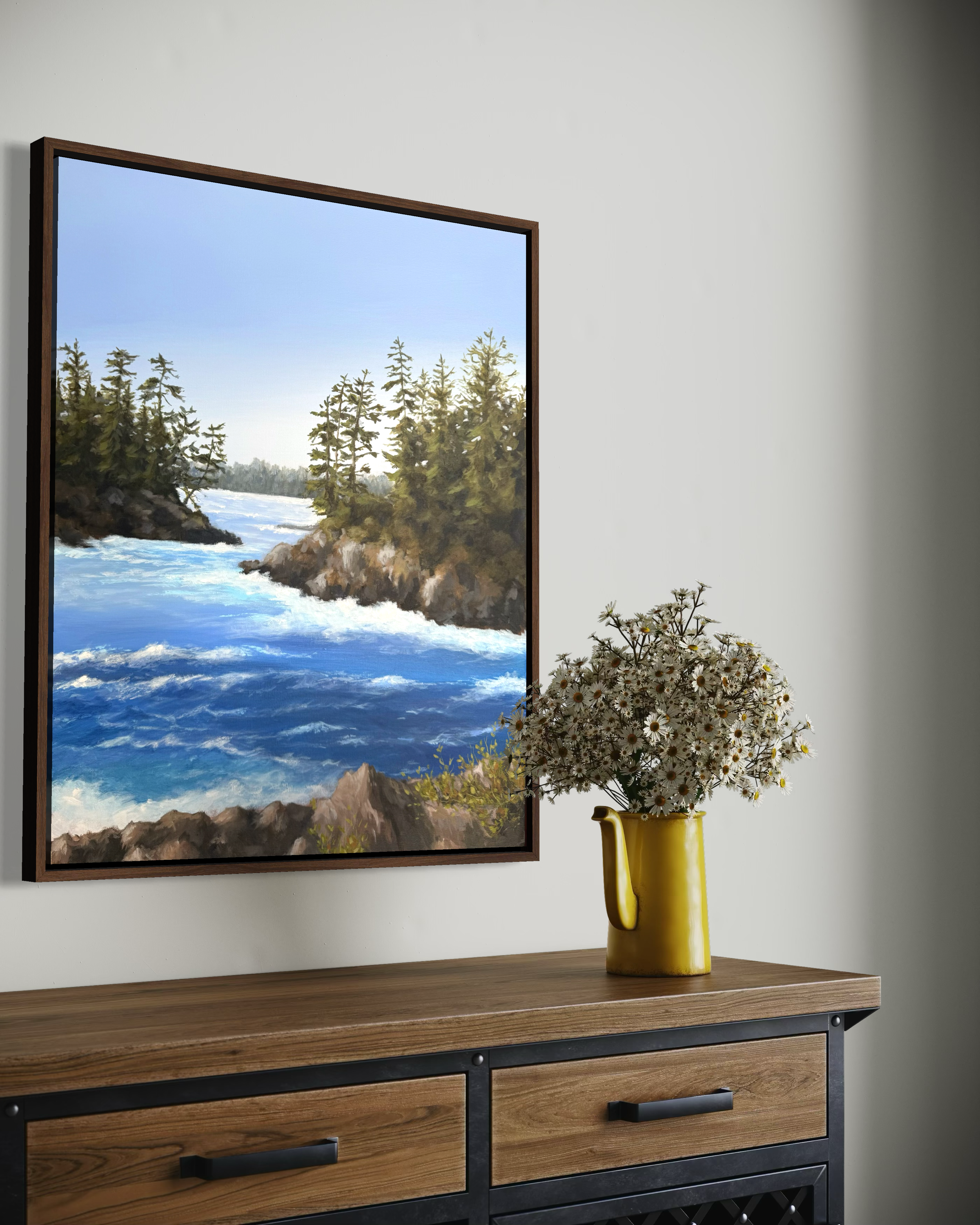 A framed landscape painting of a river with rapids, surrounded by trees on rocky cliffs, hanging on a white wall above a wooden console table with two drawers. A yellow vase filled with daisies is on the table.