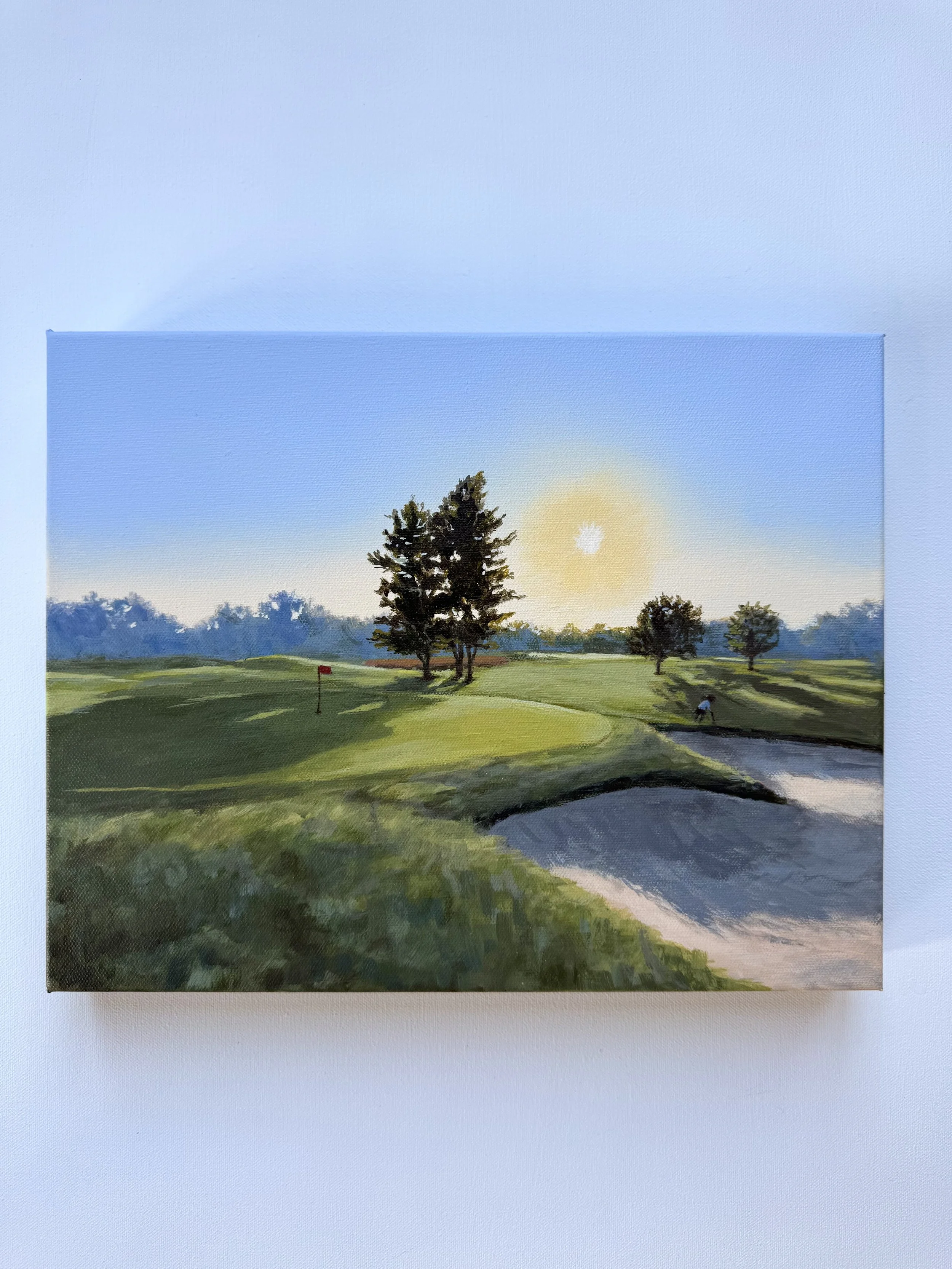 Painting of a golf course with trees, a flag, and a sand trap at sunset.