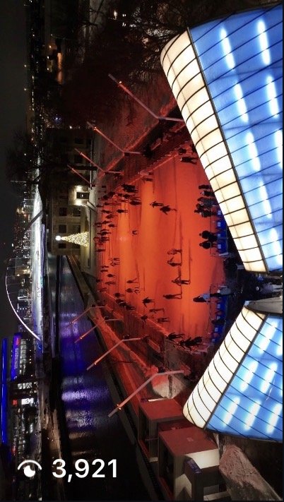Nighttime view of an outdoor ice skating rink illuminated with red and blue lights, with people skateboarding and walking around, and a modern curved glass building nearby.