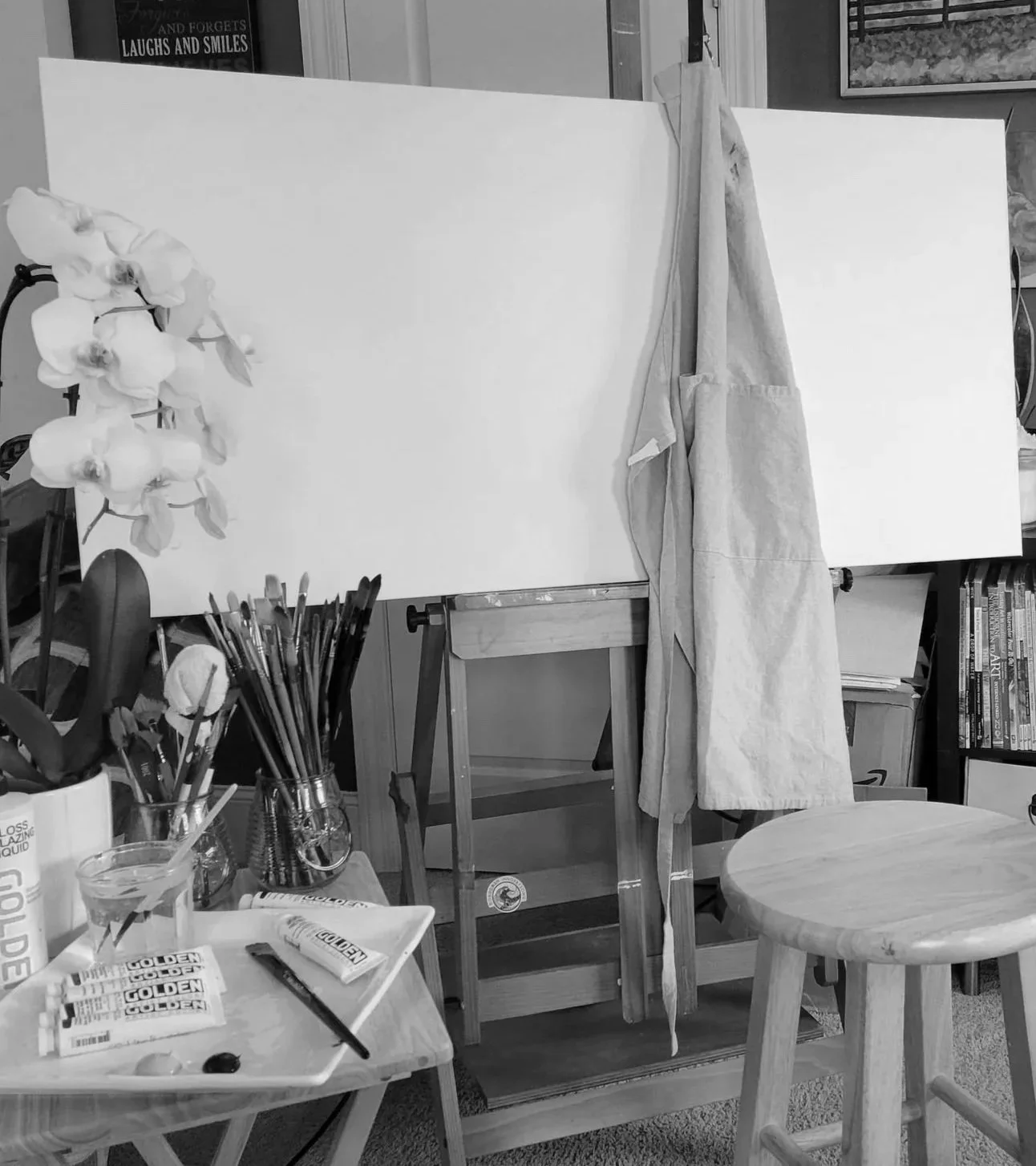 An empty canvas on an easel in an art studio, surrounded by art supplies such as brushes, paints, and a glass of water, with books and décor in the background.