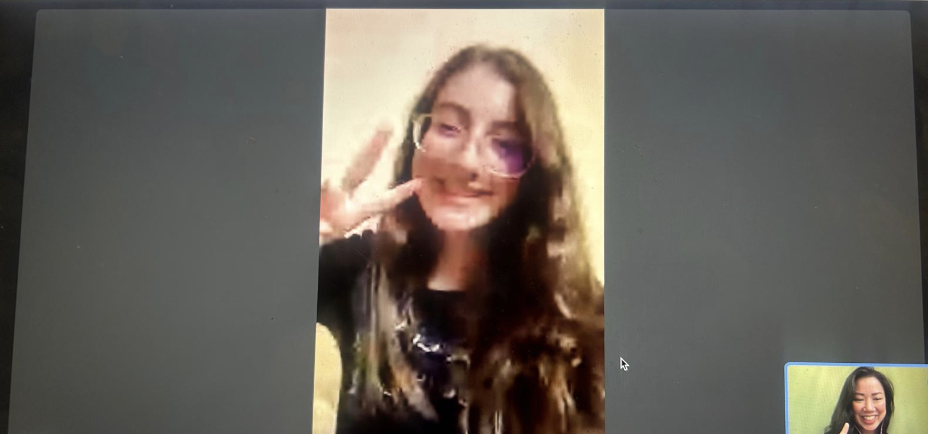 A woman with long brown hair smiling and making a peace sign with her right hand, wearing sunglasses and a black jacket in a video call.