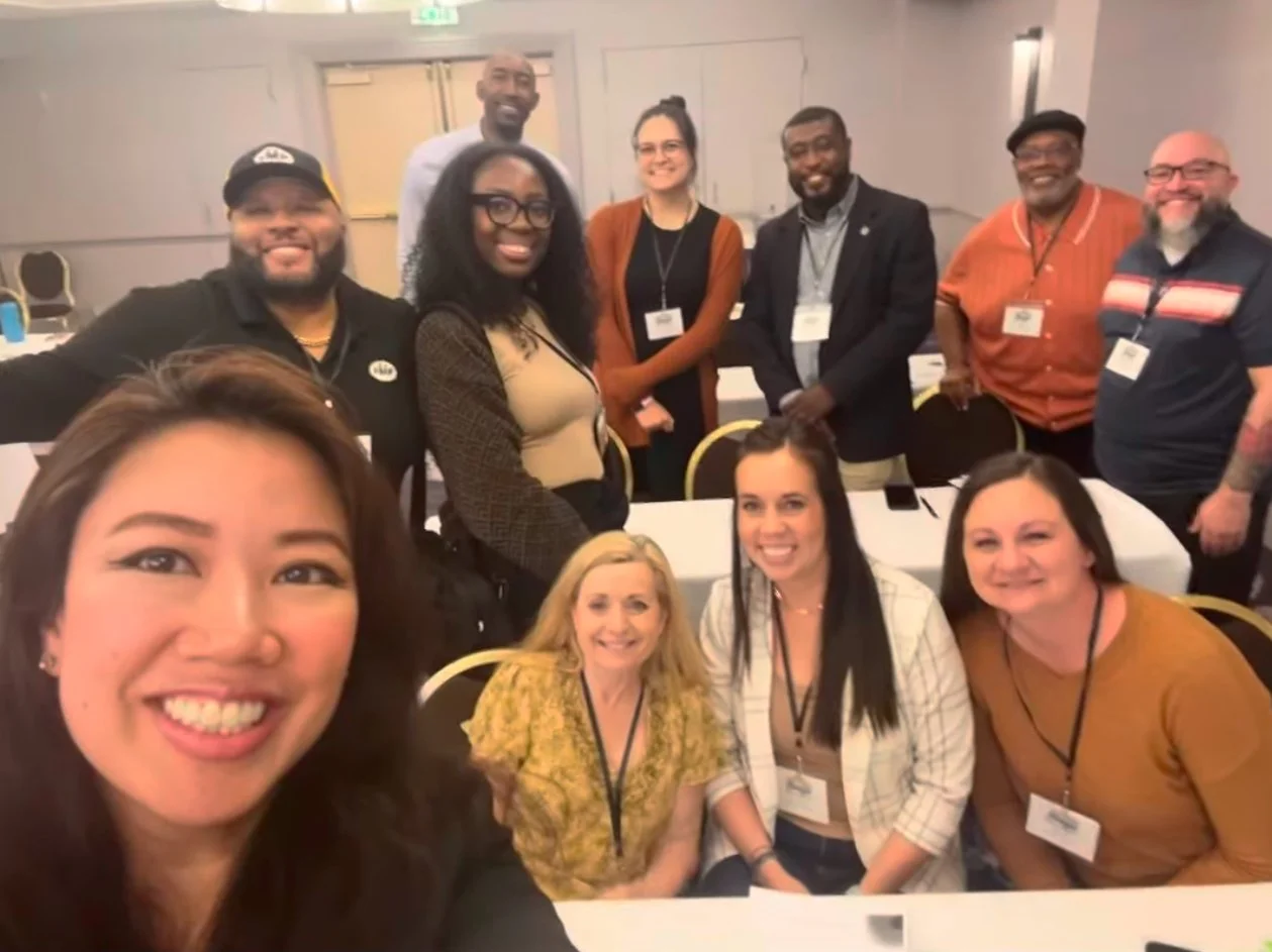Group of ten diverse people smiling at a conference, some wearing name tags, in a well-lit room with chairs around tables.
