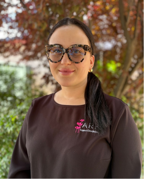 A smiling woman with long dark hair and bold torteshell rimmed glasses. She is outdoors and wears a black top with the ARK support coordinator logo on it.