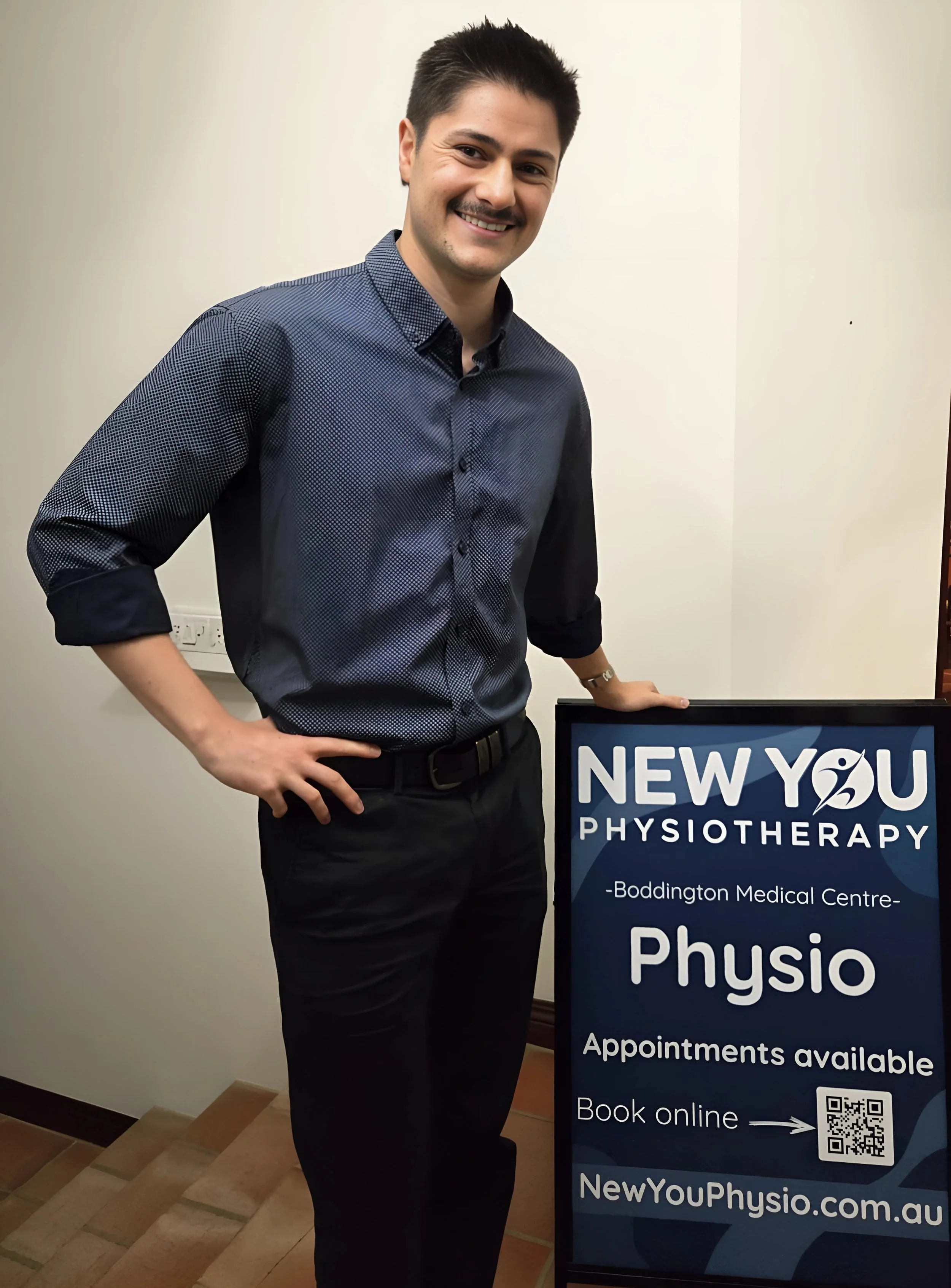 A man standing next to a sign for New You Physiotherapy, smiling, wearing a dark blue shirt and black pants, inside a building with beige walls and brown tiled floors.