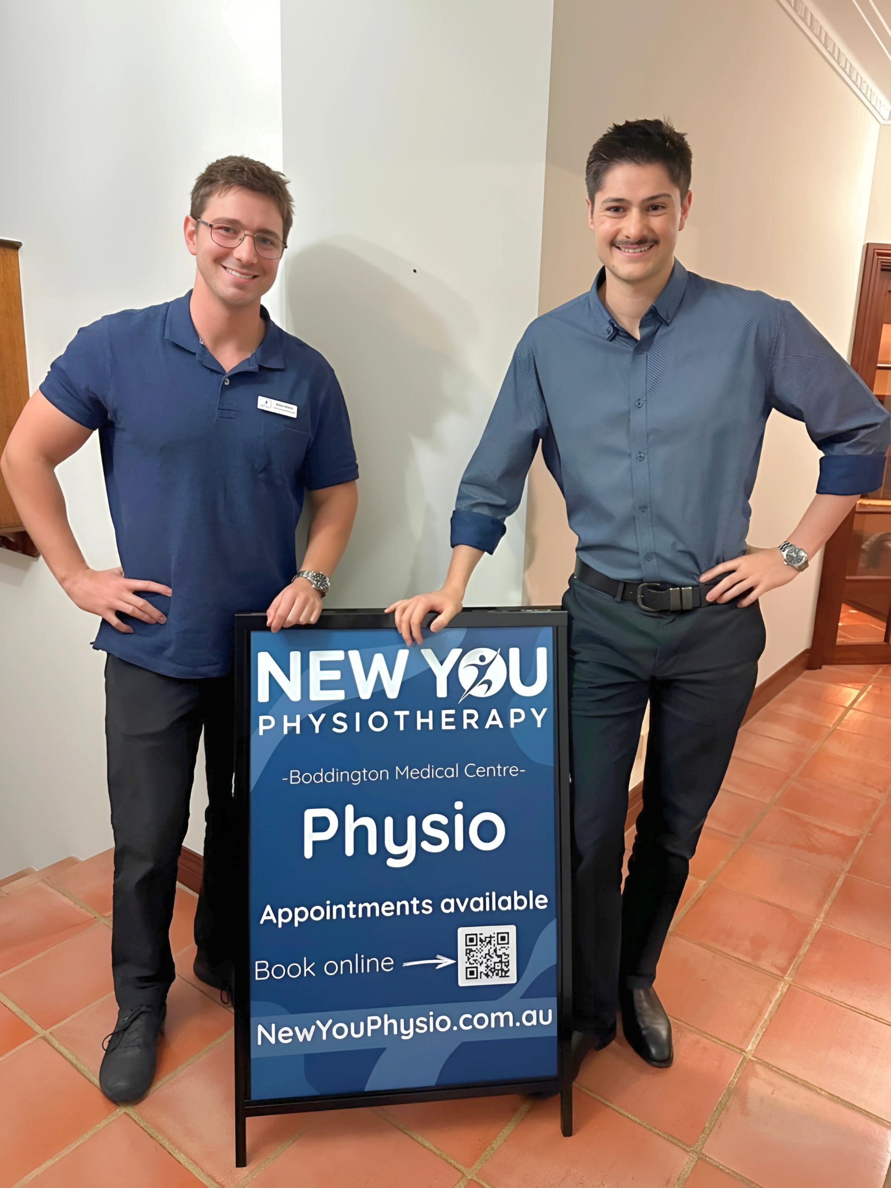 Two men standing beside a sign for New You Physiotherapy at Boddington Medical Centre, with one man in a blue polo shirt and the other in a blue dress shirt, both smiling.