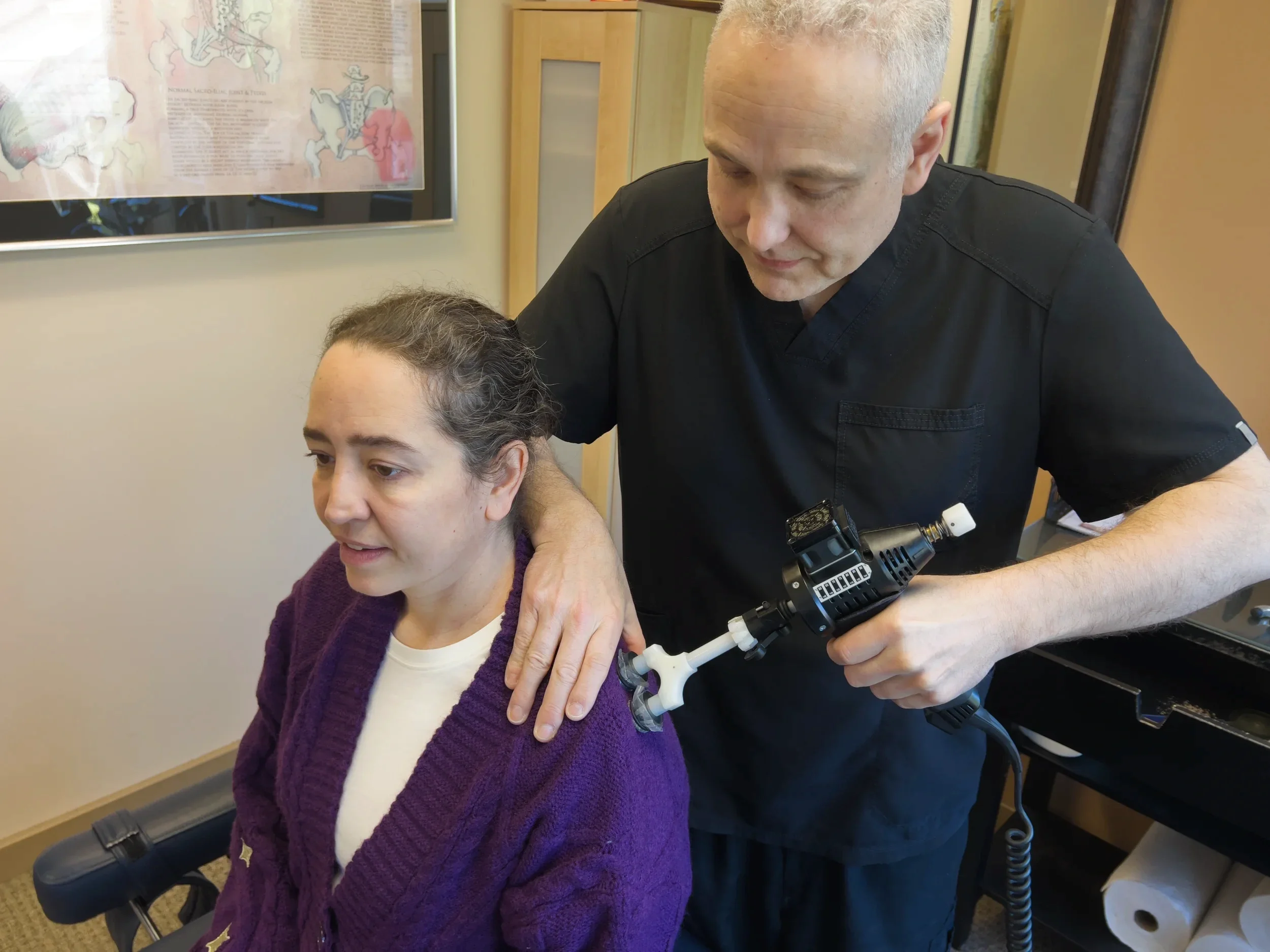 Dr. Owens in black scrubs holding an adjusting instrument on a woman's shoulder during a medical examination in a clinic.