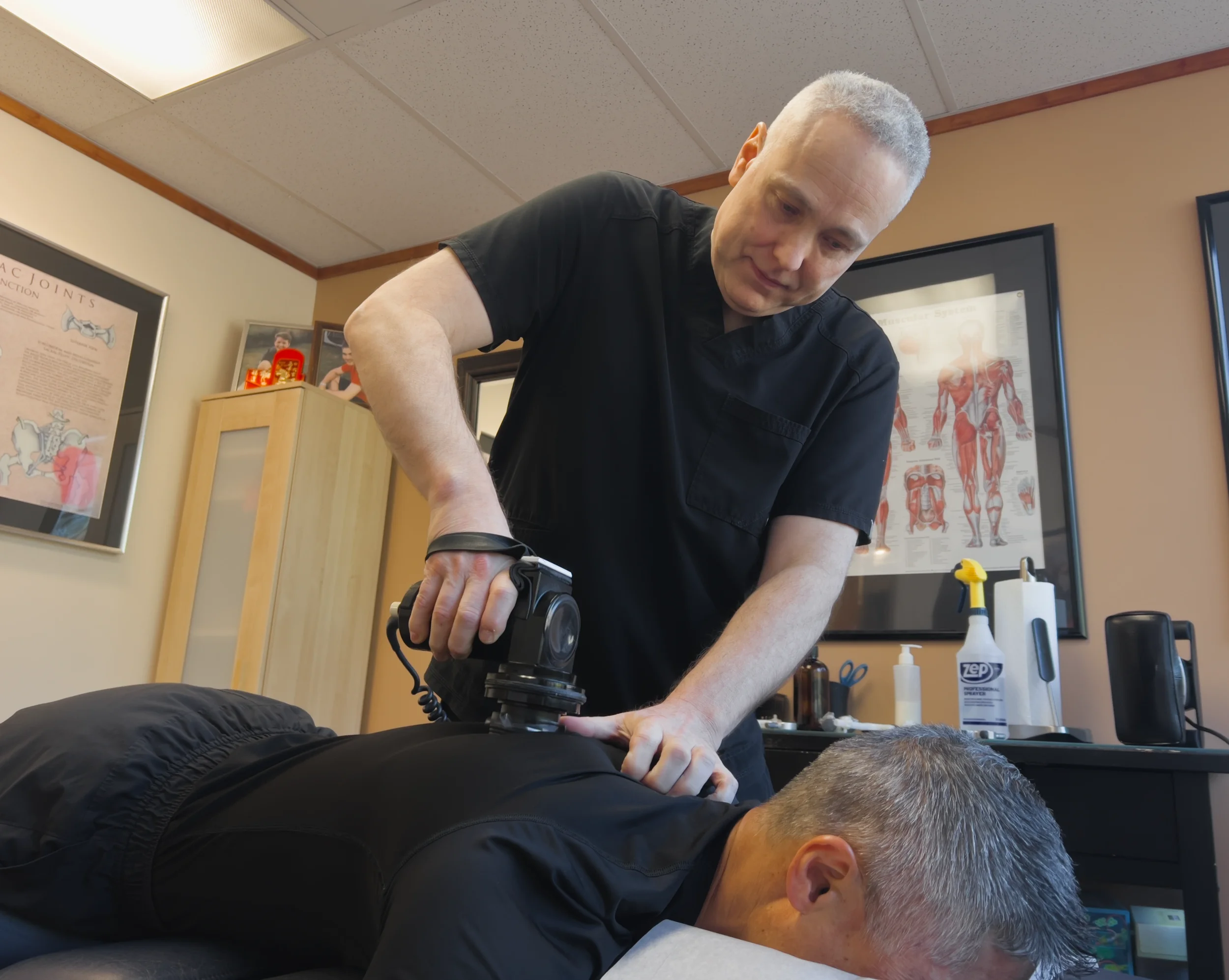 A chiropractor adjusting a patient's back on a treatment table during a therapy session in a clinic with anatomical posters on the wall.