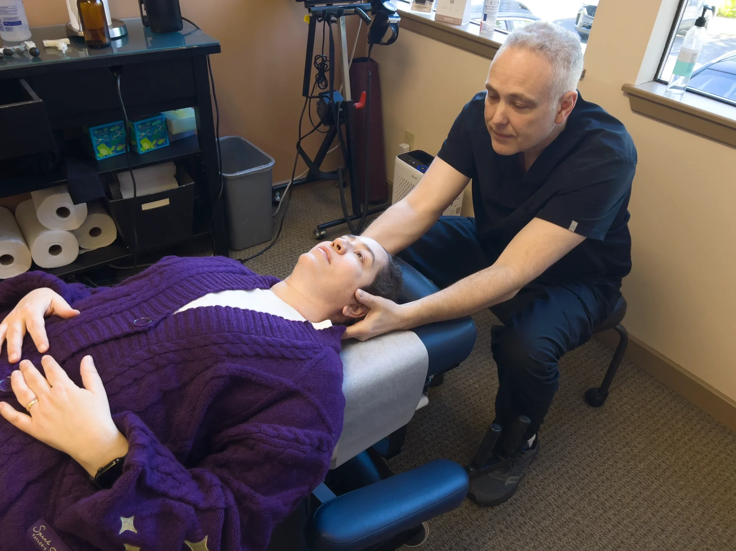 A chiropractor examines a woman lying on a treatment table, with her eyes closed, in a clinical setting with various medical supplies nearby.