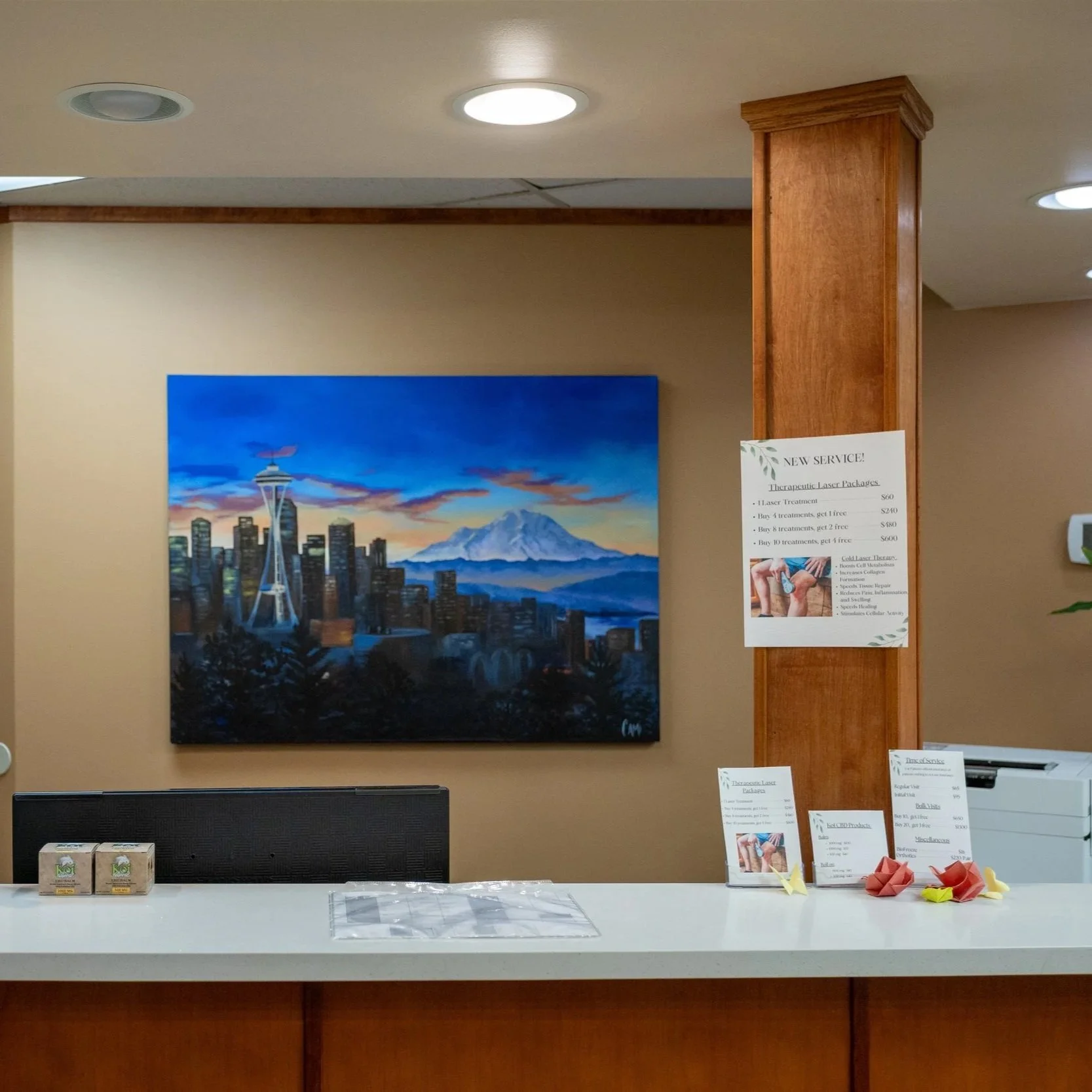 A reception counter with promotional cards and origami paper animals, a sign advertising therapeutic laser packages, and a large painting of Seattle's skyline with Mount Rainier in the background, on a beige wall with ceiling lights.