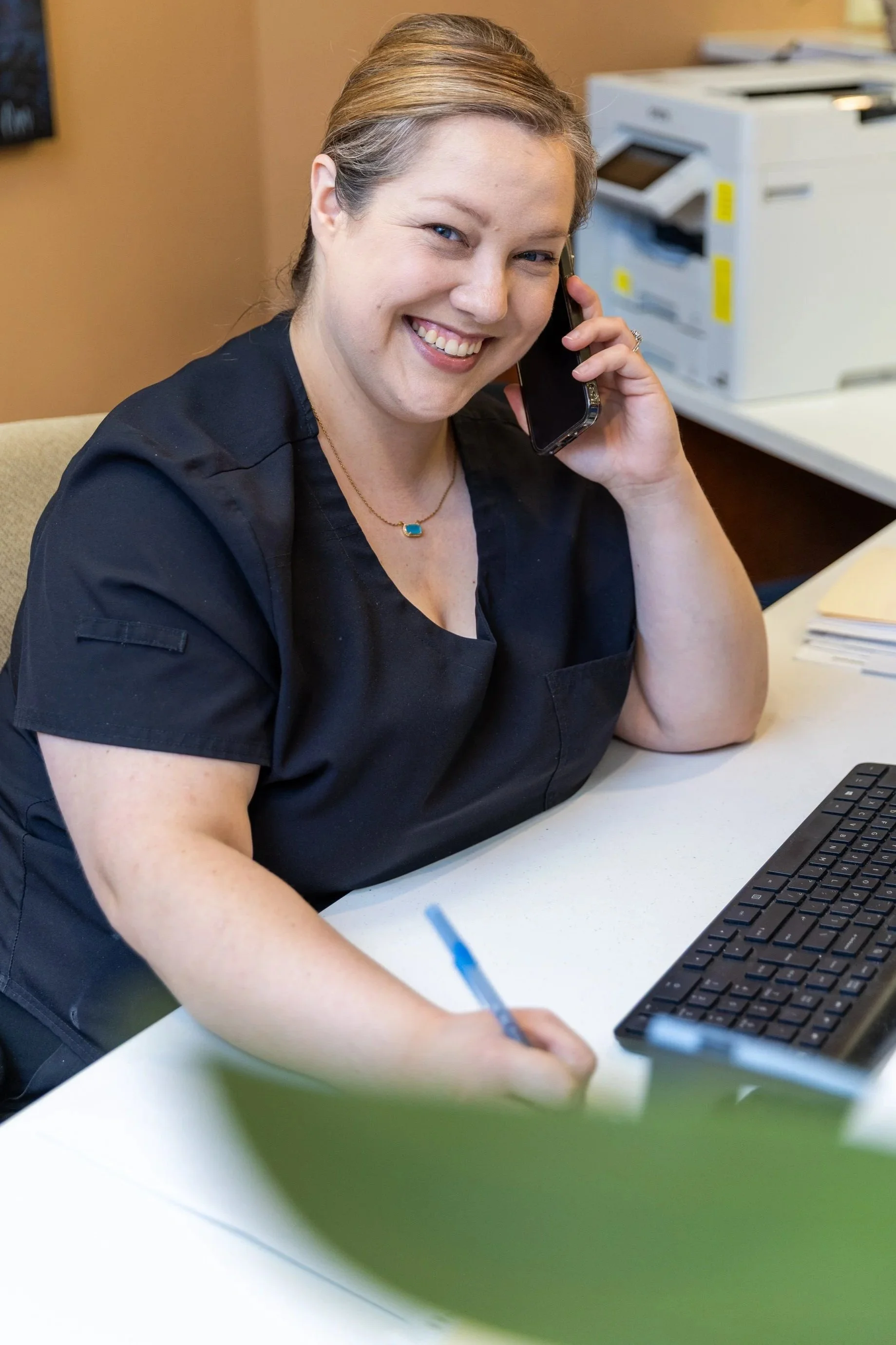 A woman smiling and talking on a mobile phone at the front desk with a computer keyboard in front of her, in an office setting.