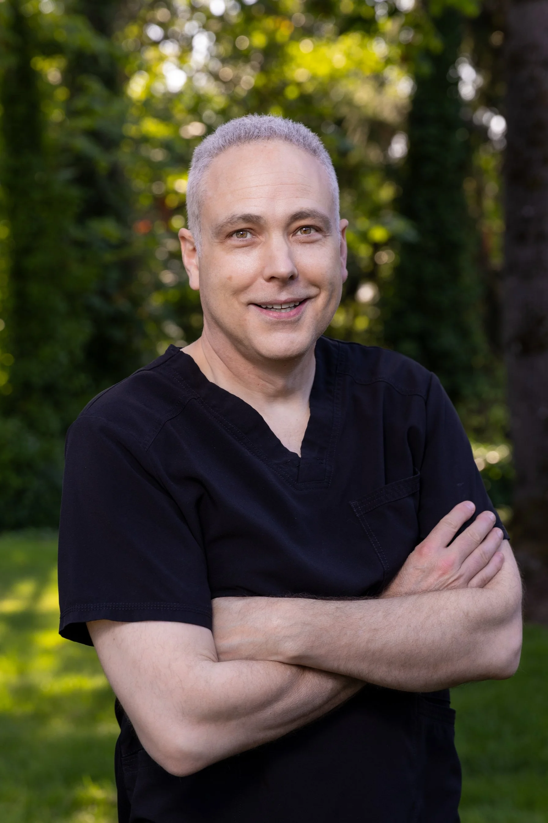 Dr. Edward Owens, wearing a black shirt, standing with his arms crossed outdoors in a lush green park with trees and sunlight filtering through the foliage.