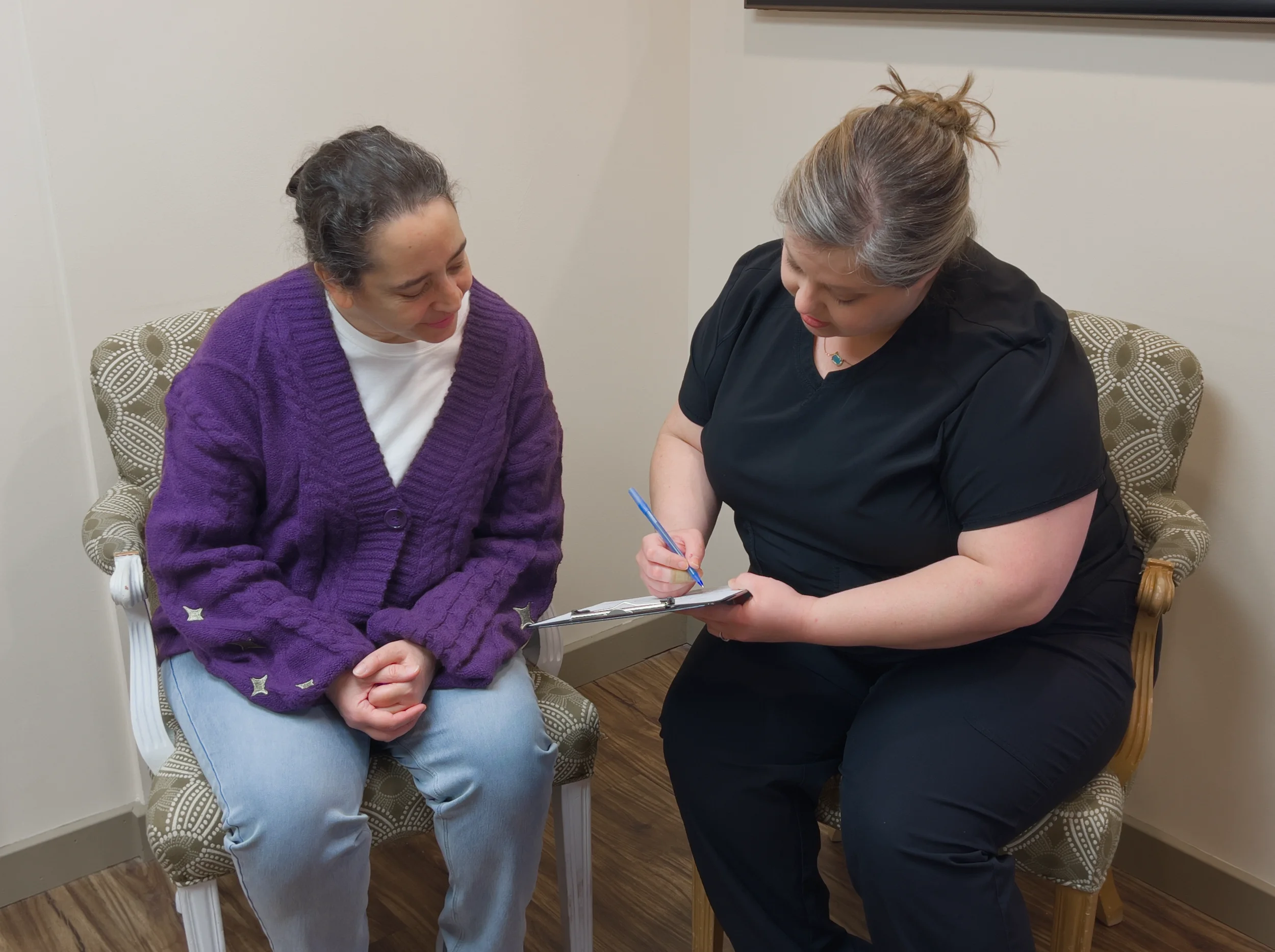 Two women sitting in chairs, one in a purple sweater and the other in black scrubs, engaged in a conversation with the woman in black taking notes on a clipboard.