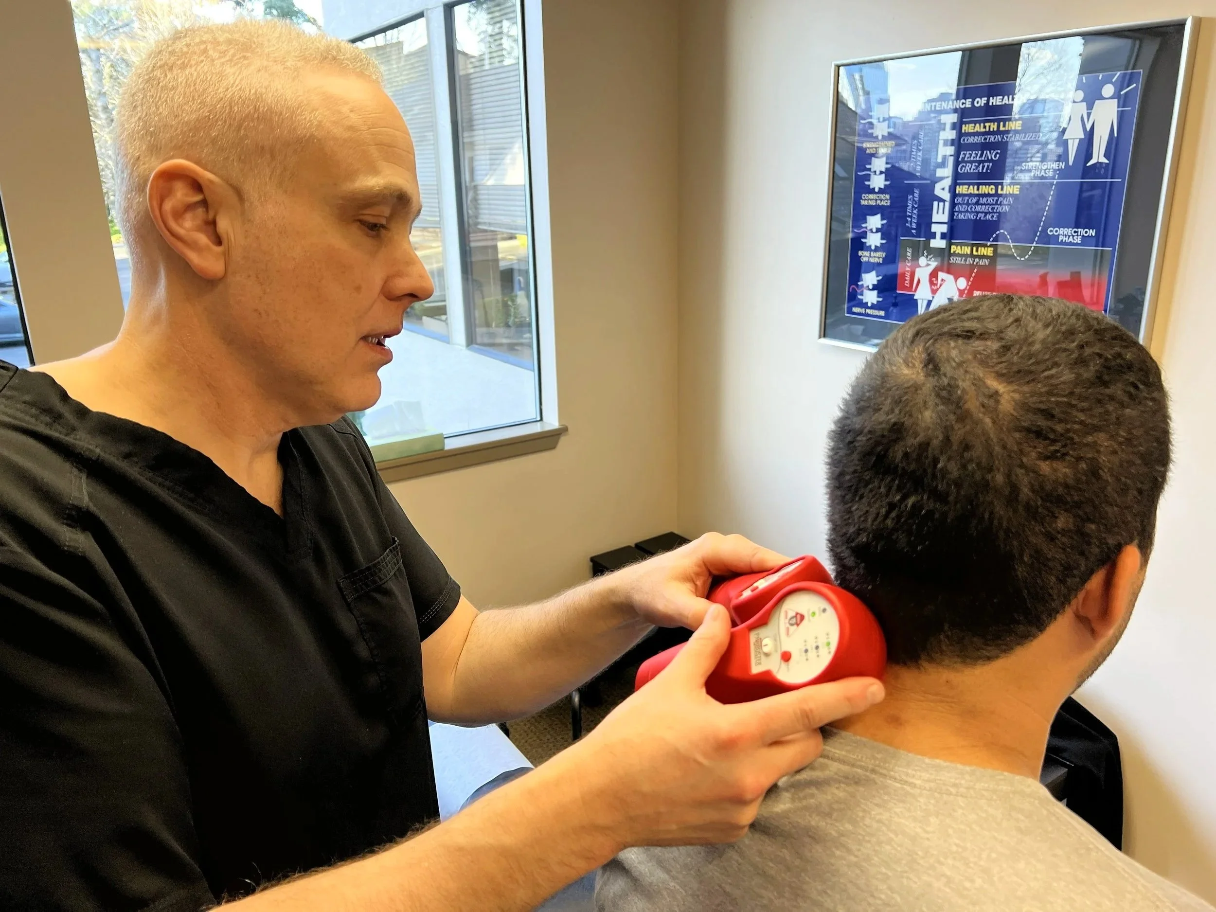 A chiropractor using a cold laser therapeutic device on a patient's neck in a clinic room, near a window and a health information poster on the wall.