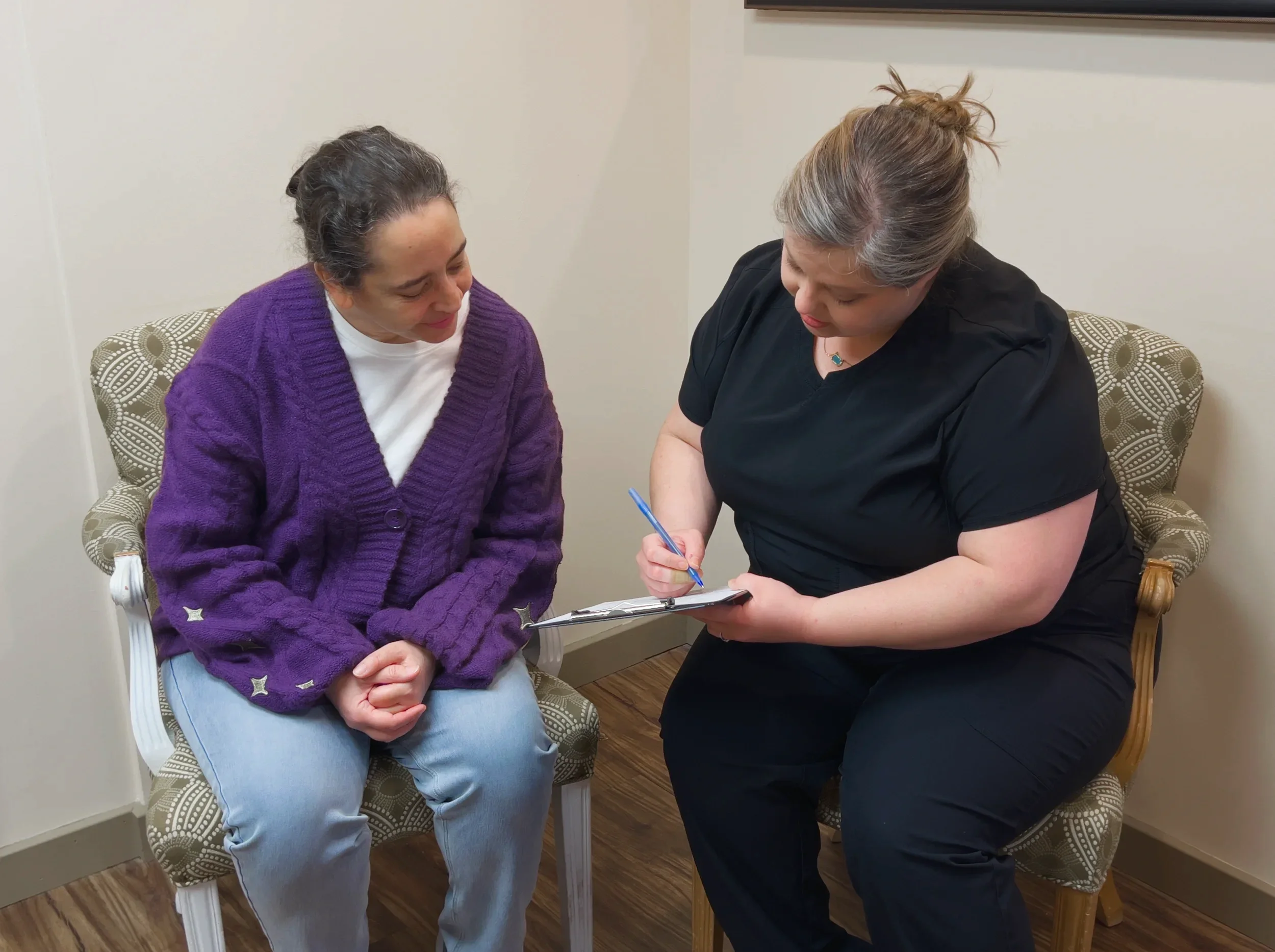 Two women sitting on chairs, one wearing a purple sweater and the other in medical scrubs, are engaged in a discussion with a clipboard and pen.